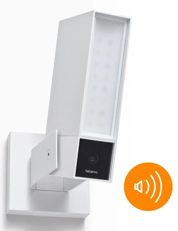 Białe urządzenie smart home Netatmo. Prostokątny korpus z ikoną głośnika. Widoczna kamera i światło.