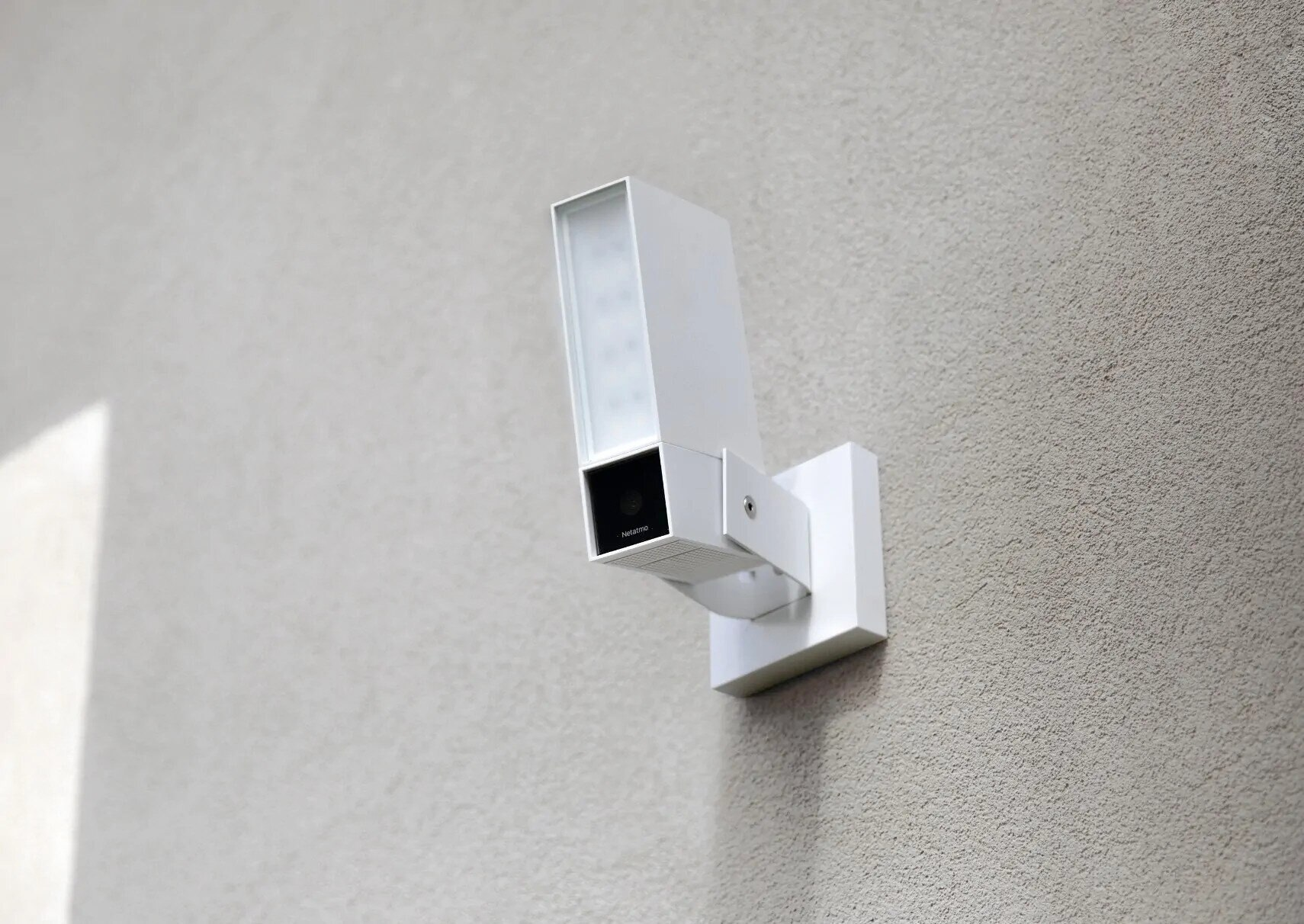 Biała kamera bezpieczeństwa zamontowana na jasno szarej ścianie. Jest prostokątna.