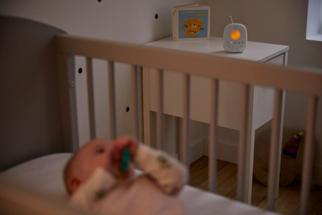 Baba a kiságyban cumival, monitorral és könyvvel egy éjjeliszekrényen.