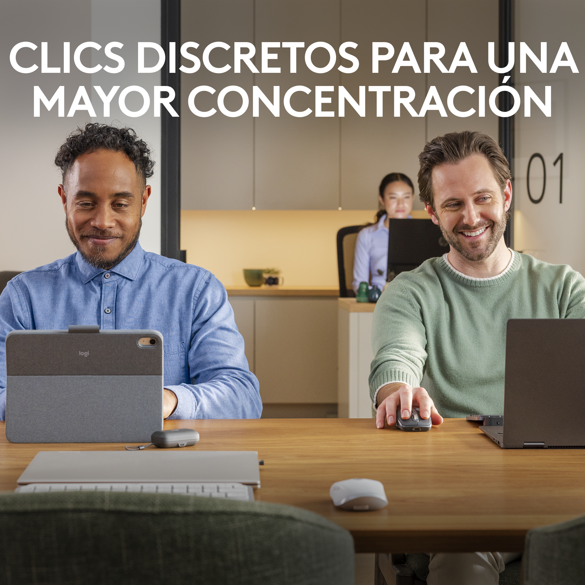 Dos hombres en una mesa usando portátiles. Uno usa un ratón. Una mujer al fondo. El esquema de color es claro.