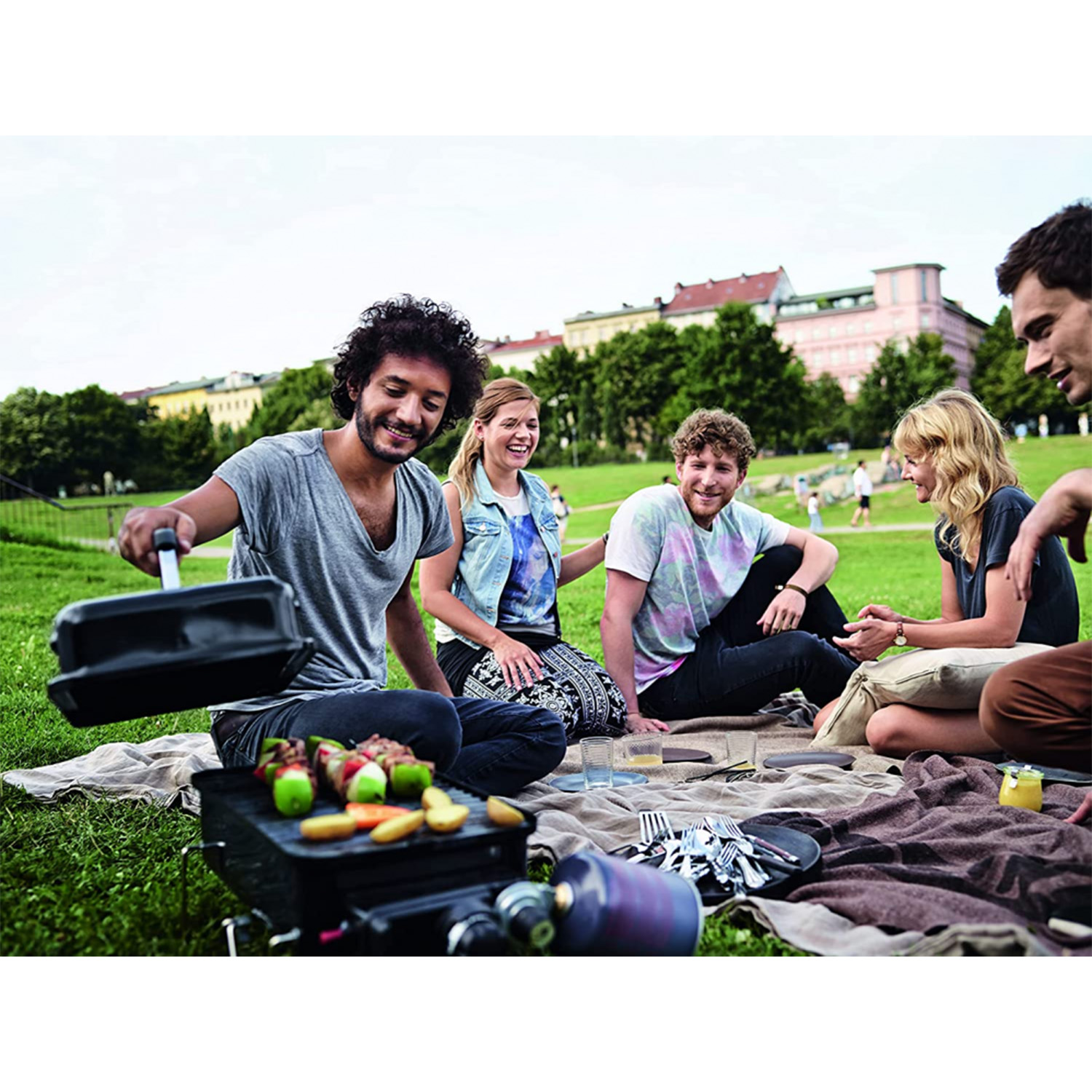 Gruppo di persone che grigliano cibo su una piccola griglia in un parco, con alberi ed edifici sullo sfondo.