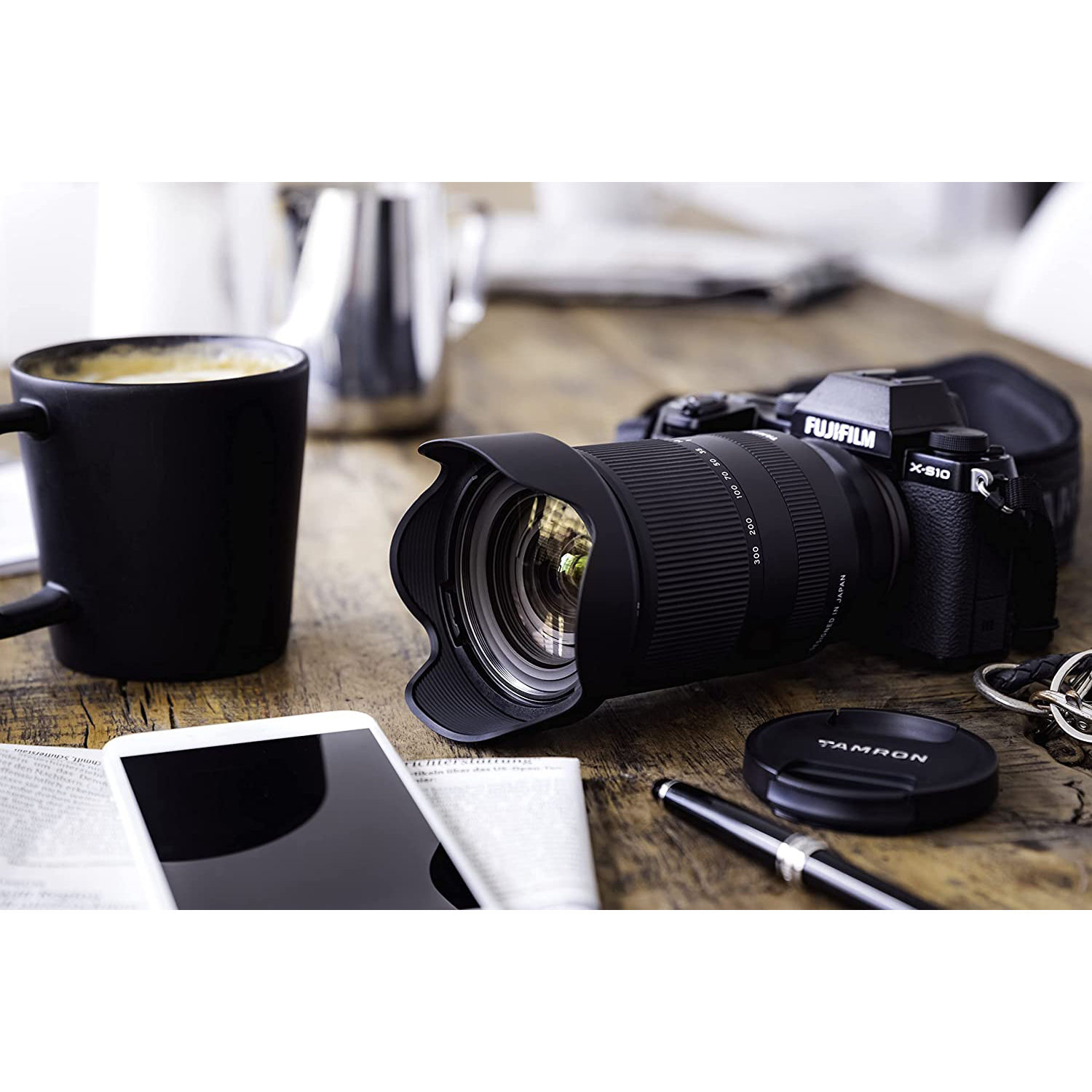 Fotocamera con obiettivo, tazza di caffè, telefono e chiavi su tavolo di legno.