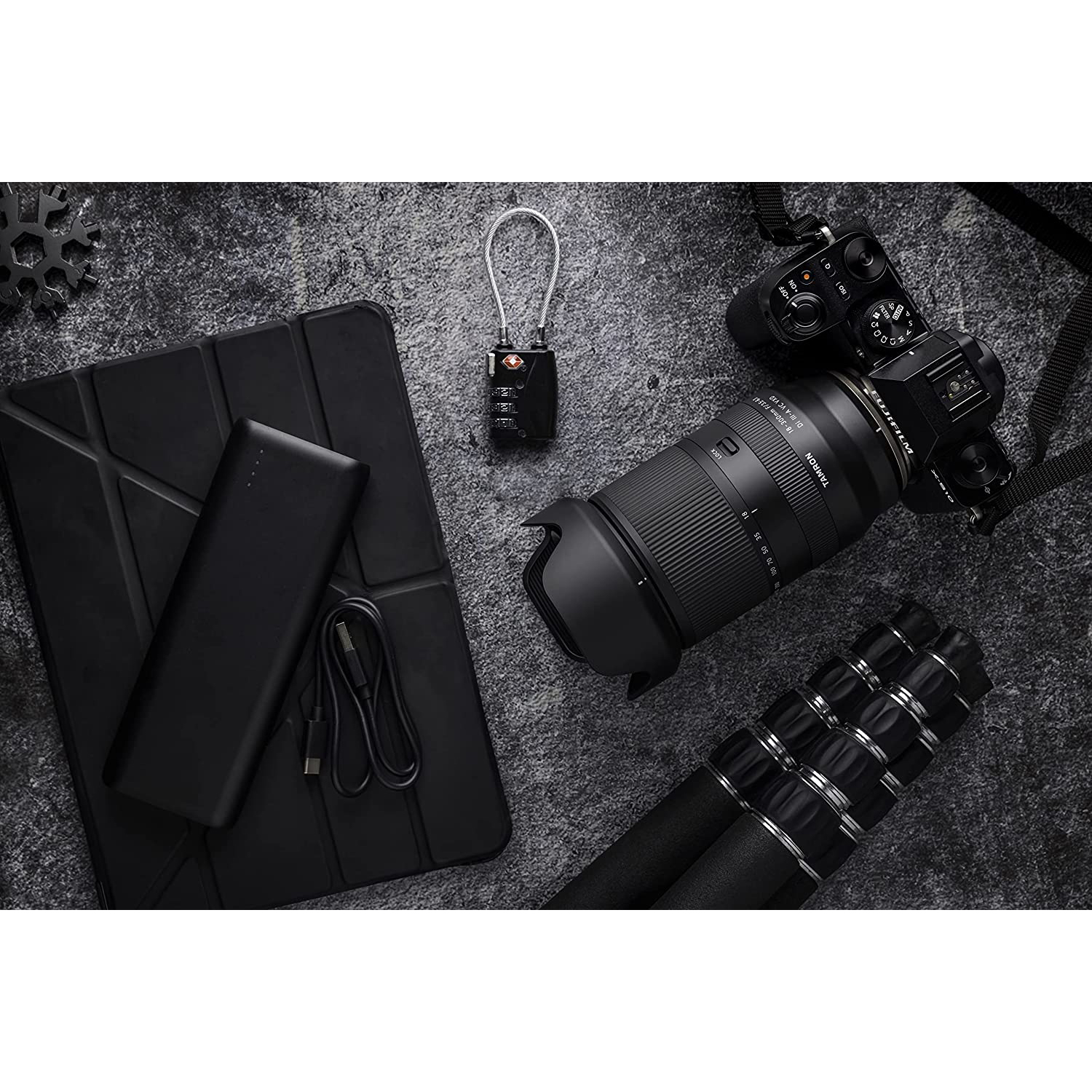 Accessori neri: tablet, lucchetto, fotocamera con obiettivo, treppiede, cavo USB, su superficie strutturata.