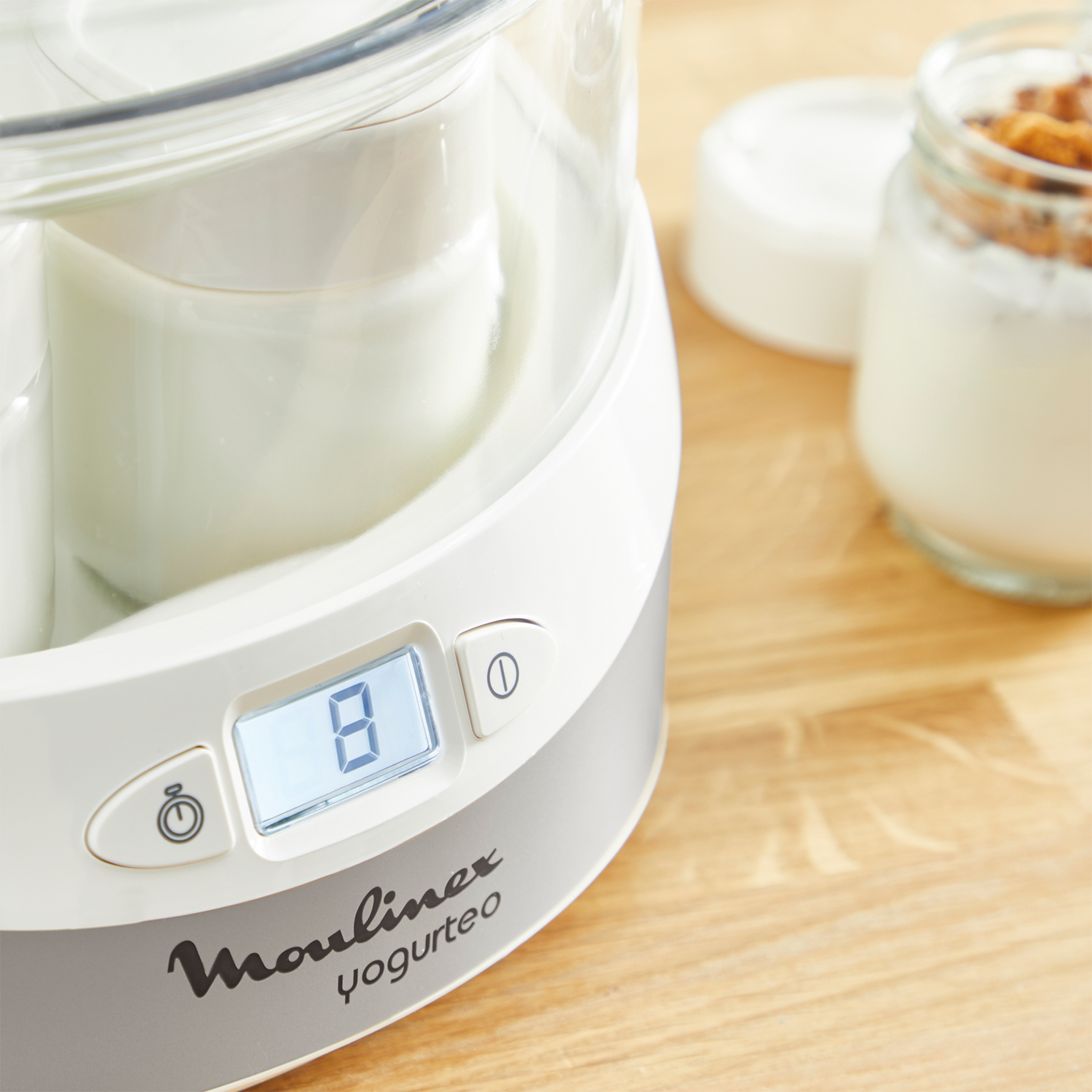 Una yogurtiera bianca e grigia con display digitale che mostra il numero 8. Un vasetto di yogurt a destra.
