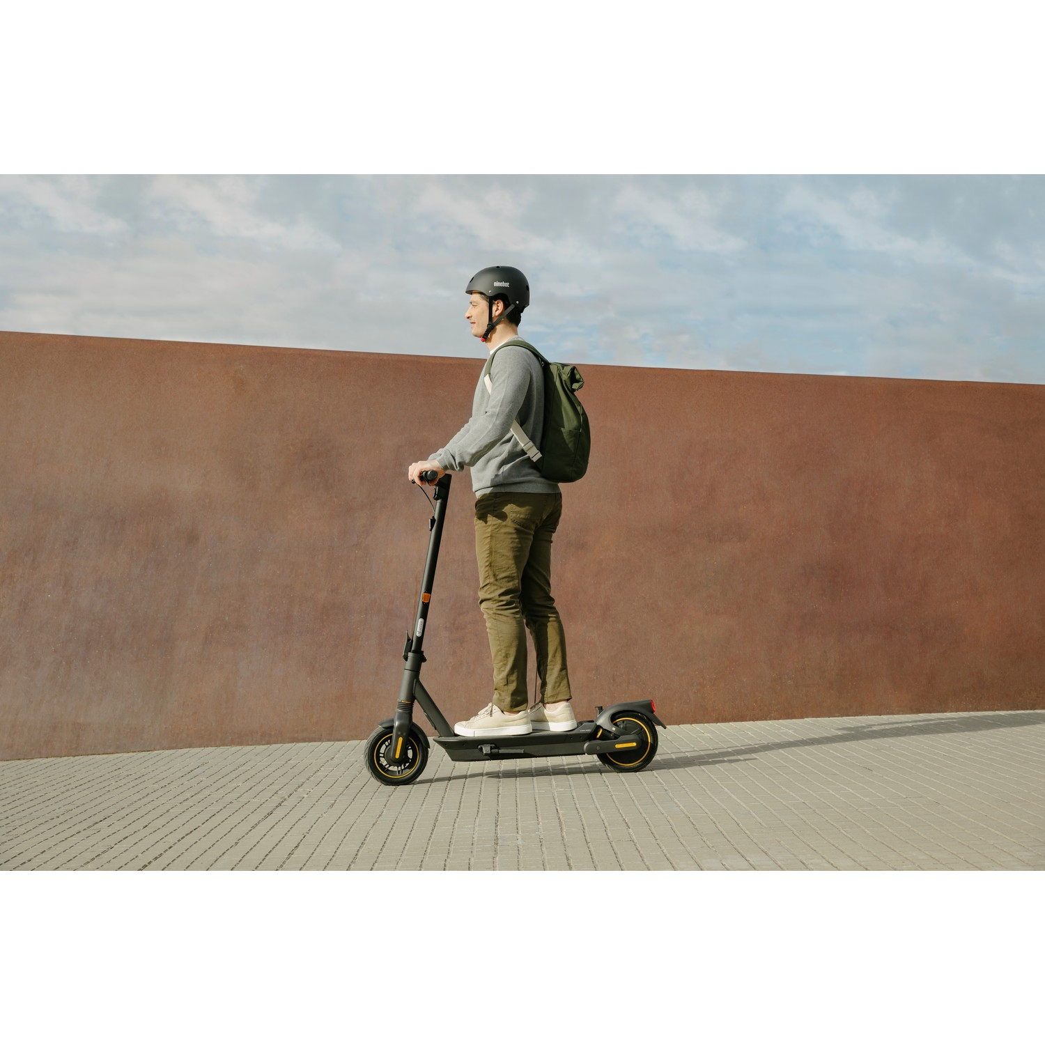 Un homme conduisant une trottinette électrique noire et jaune sur un chemin de briques, avec un mur marron.