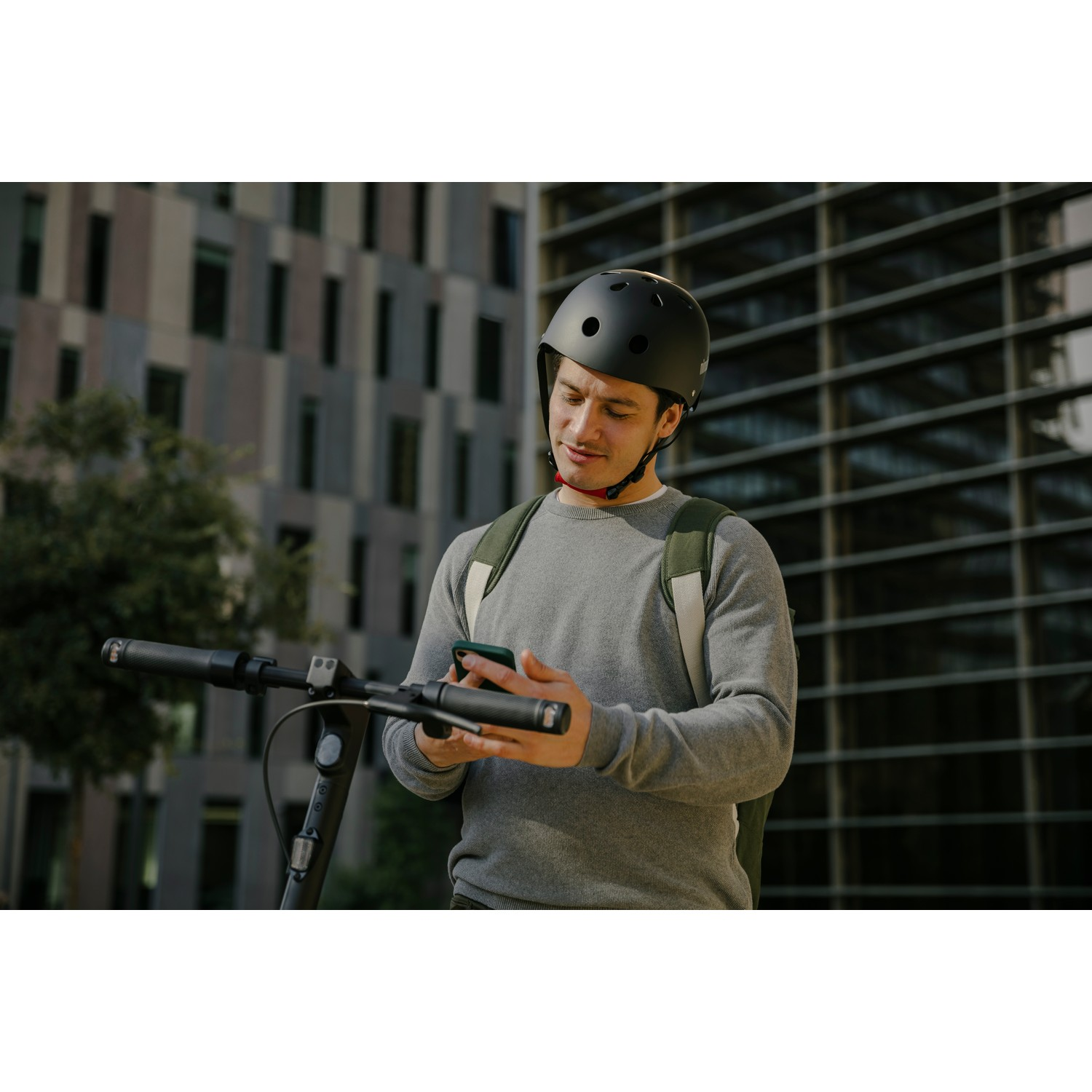Homme en casque utilisant son téléphone près d'un scooter. Bâtiments et arbres en arrière-plan.