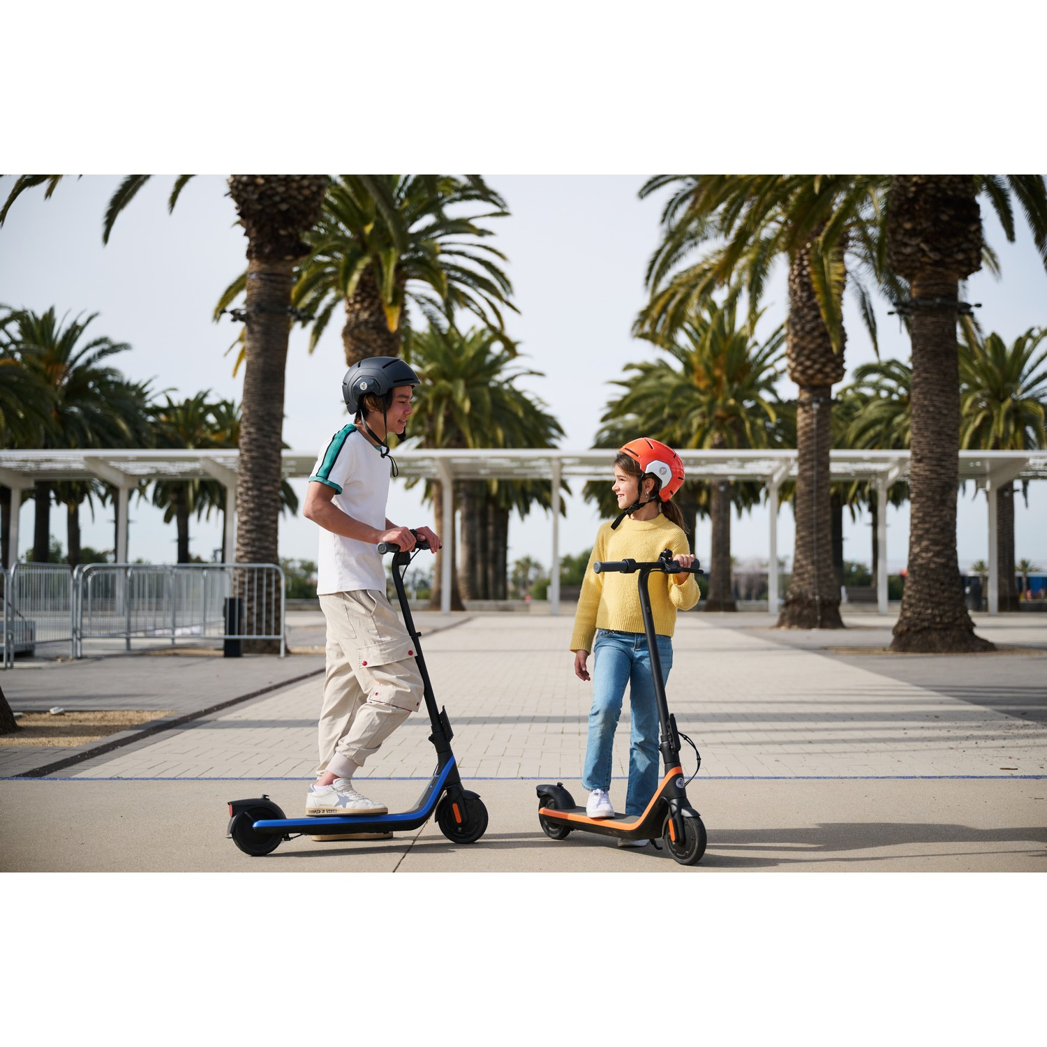 Two children on scooters. One wears a black helmet and the other an orange helmet. Palm trees are behind them.