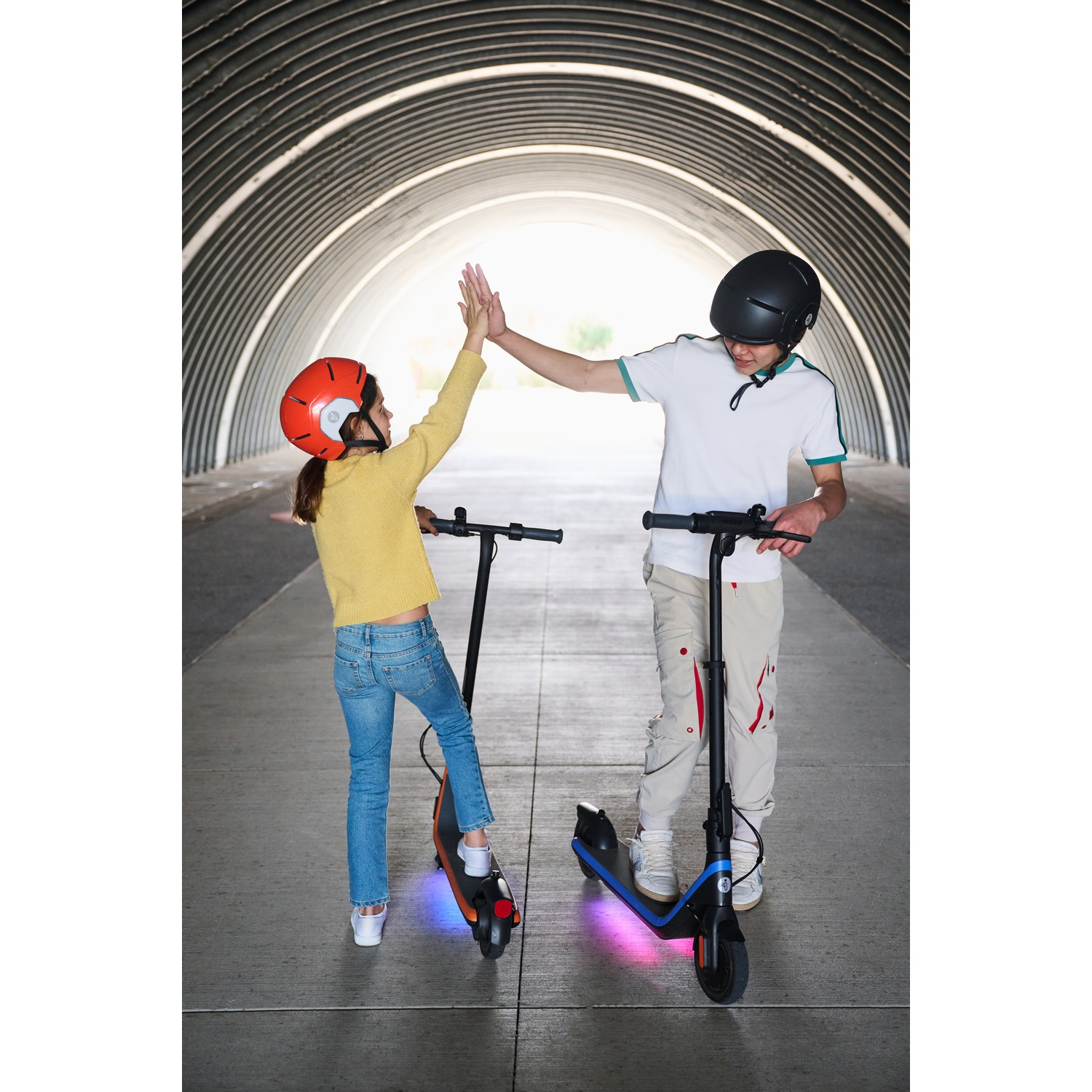 Twee kinderen op scooters geven elkaar een high-five in een tunnel. Eén heeft een rode helm.