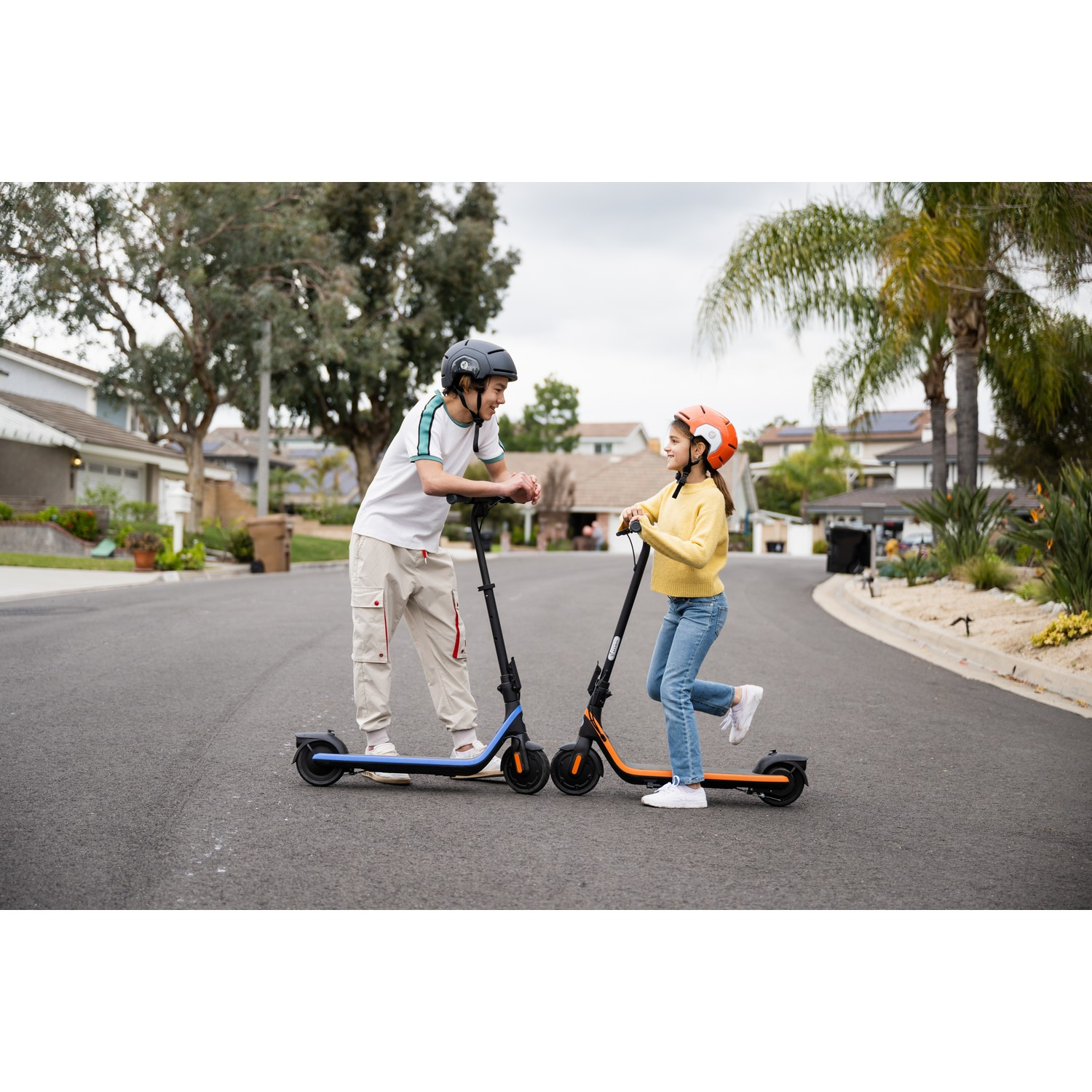 Twee kinderen op scooters, één blauw, één oranje, op een straat in een buitenwijk.