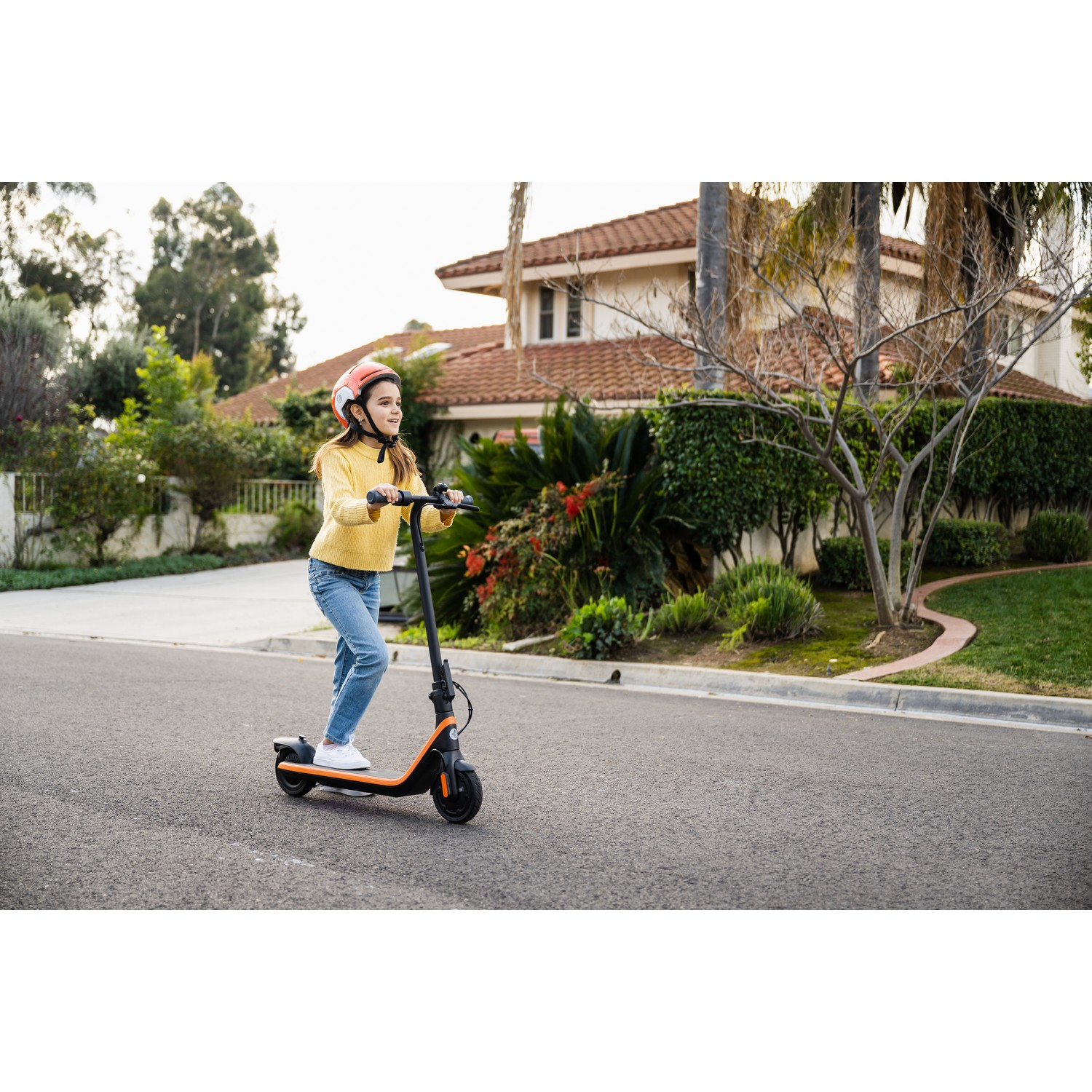 Meisje op een scooter op een weg. Draagt een helm. Huizen op de achtergrond.