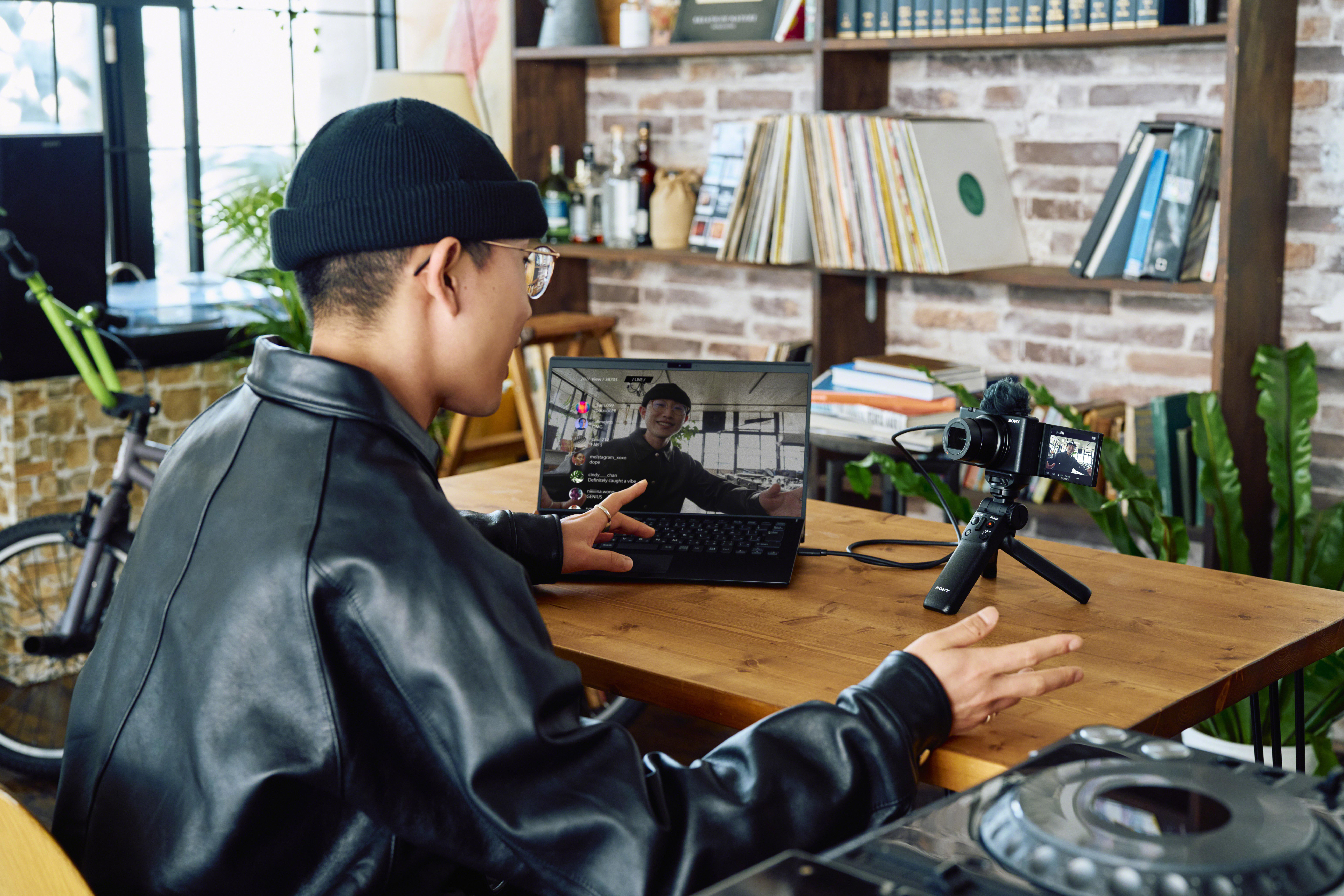 Ein Mann in einer schwarzen Mütze und Jacke führt eine Videokonferenz auf einem Laptop durch. Eine Kamera und ein Mischpult sind zu sehen.