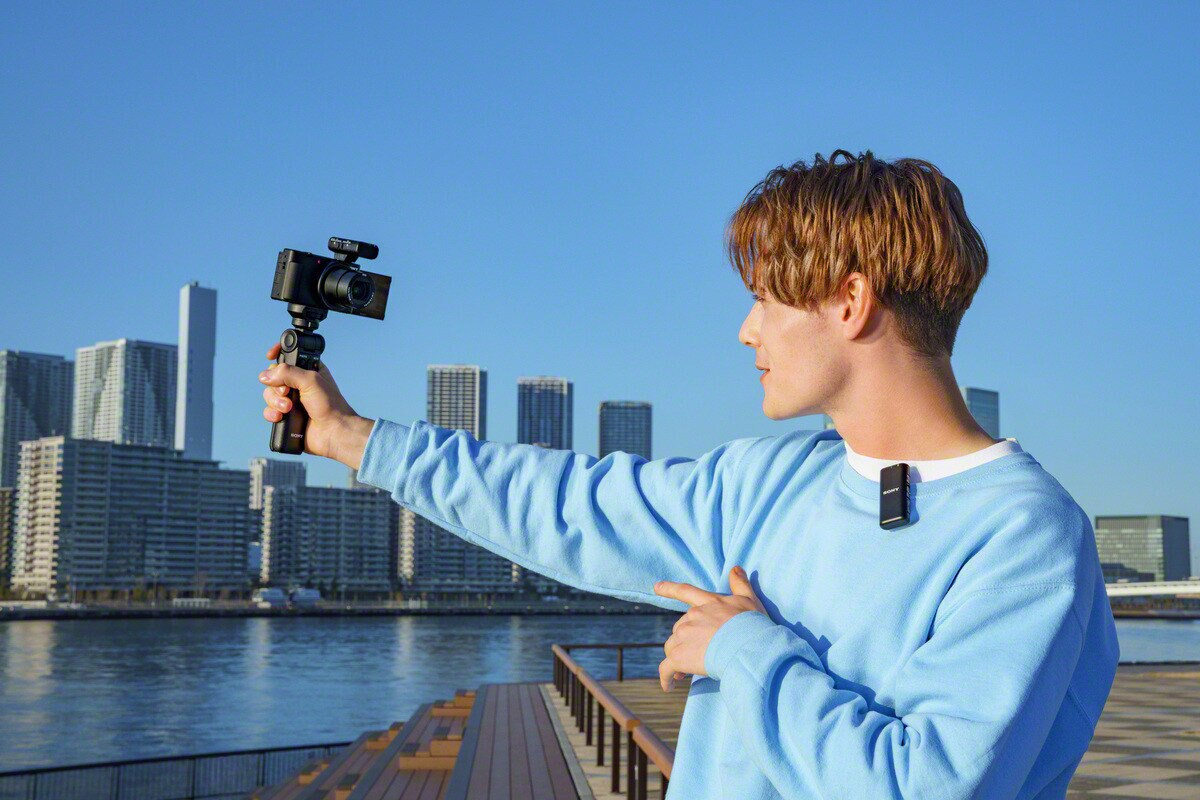 Mężczyzna trzymający kamerę na małym statywie, mówiący do czarnego mikrofonu, z tłem w postaci krajobrazu miejskiego.