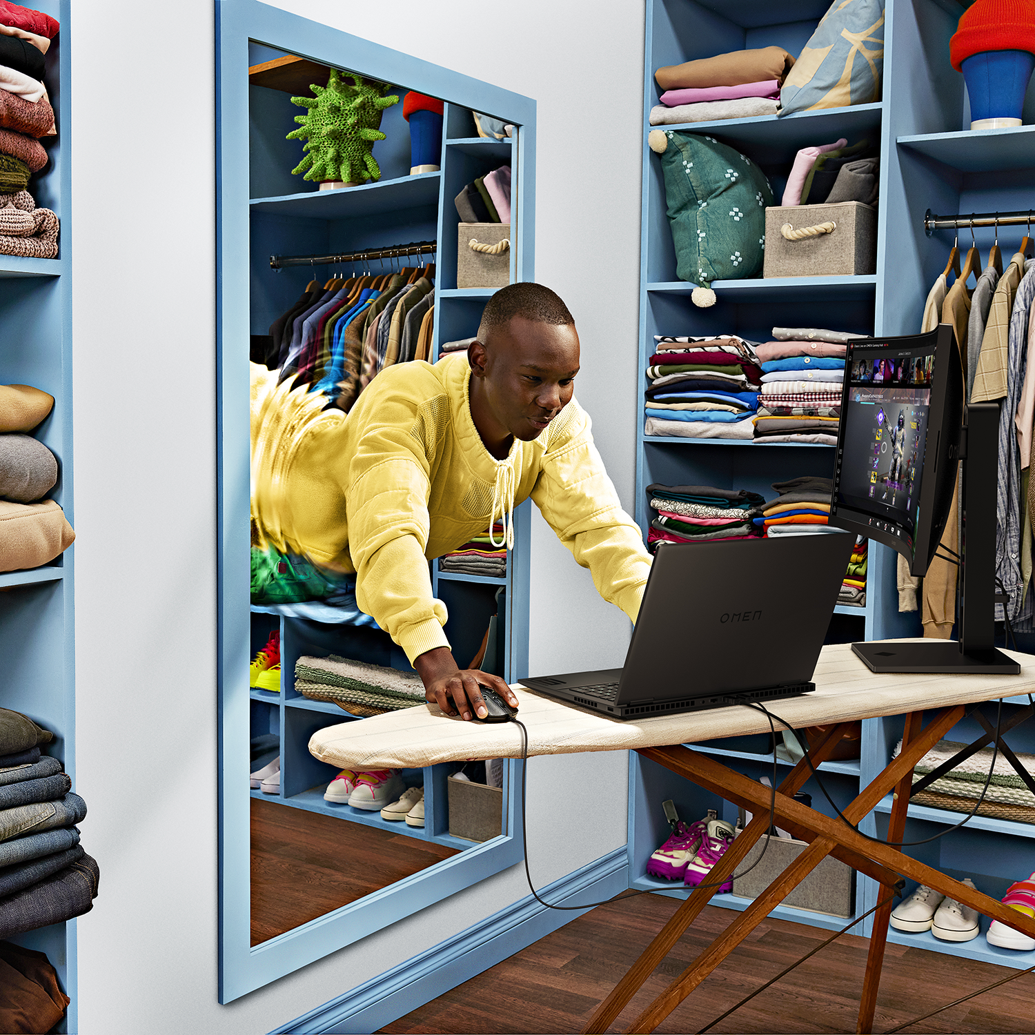 Man using a laptop on an ironing board, reflected in a mirror in a blue closet. A monitor is present.