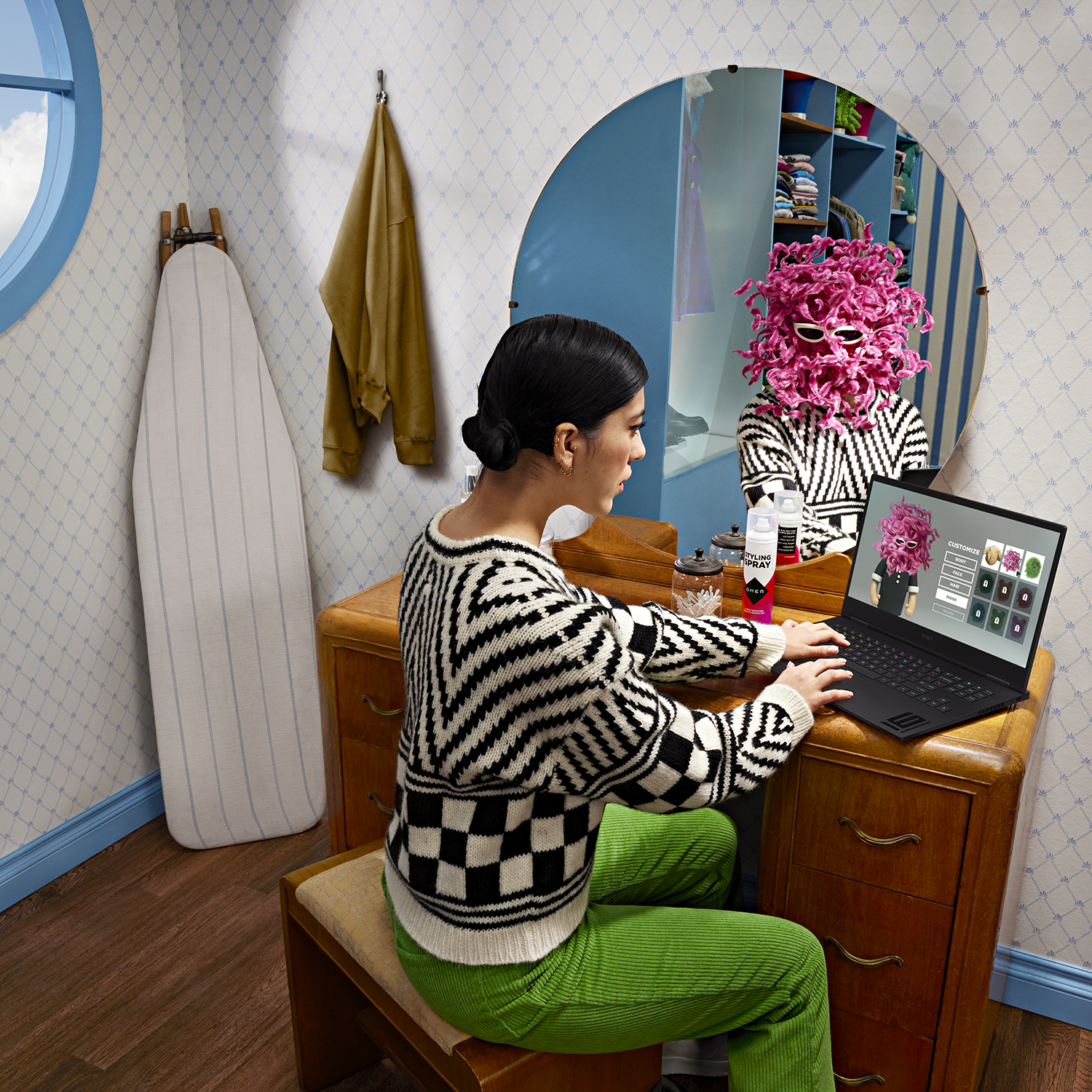 A woman sits at a vanity table looking in the mirror, with a laptop in front of her.