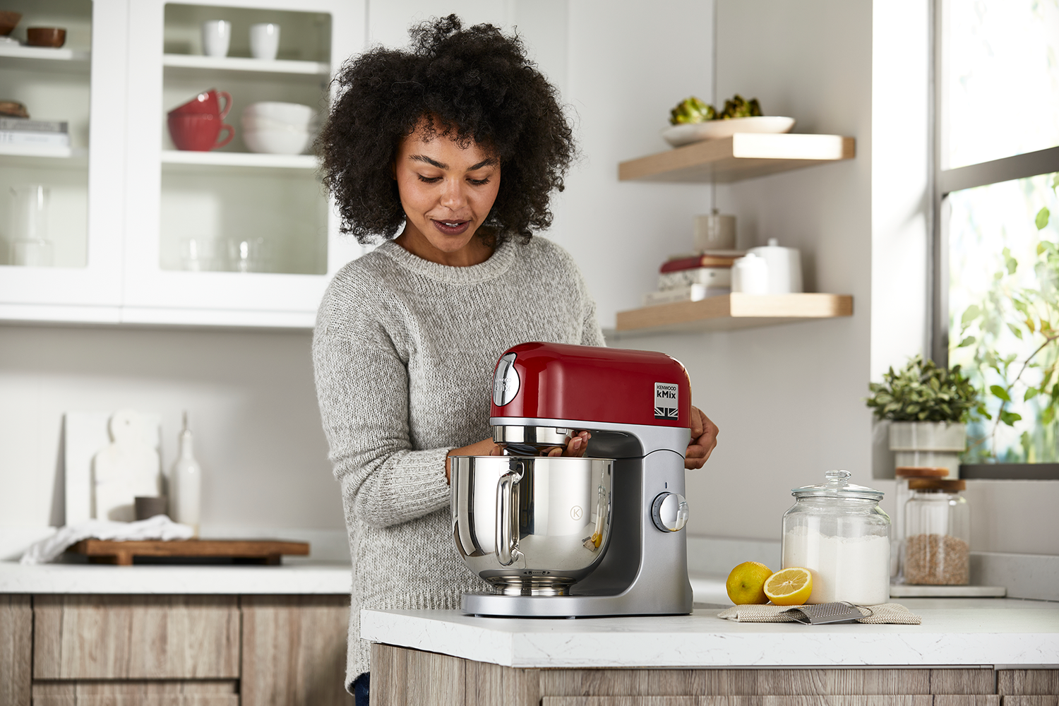 Een vrouw die een rode en zilveren keukenmachine gebruikt in een keuken. Een pot en citroenen staan in de buurt.