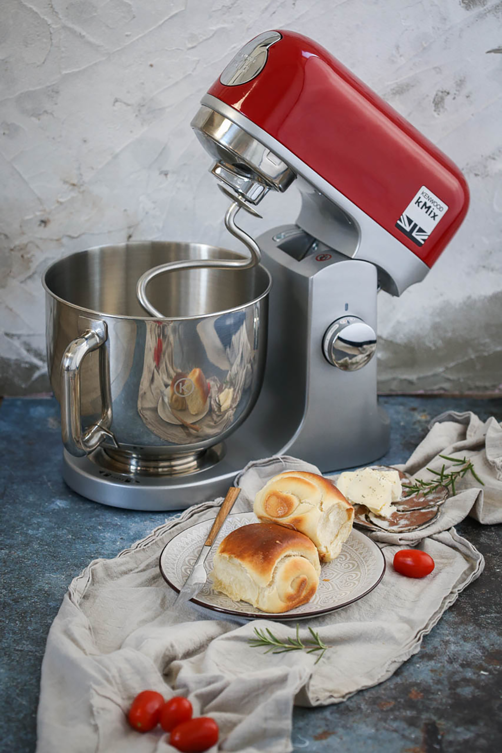Een rode Kenwood mixer met een stalen kom. Broodjes en tomaten liggen op tafel.