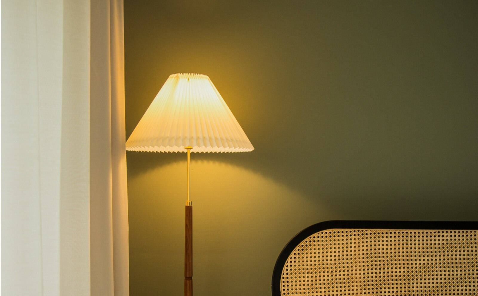 Lampa podłogowa z białym plisowanym kloszem, drewnianą podstawą i ciemnozieloną ścianą w tle.
