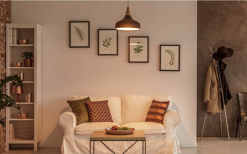 Salon z sofą, grafikami i lampą. Widoczny wieszak i regał. Ściany są neutralne z sekcją ceglaną i betonową.