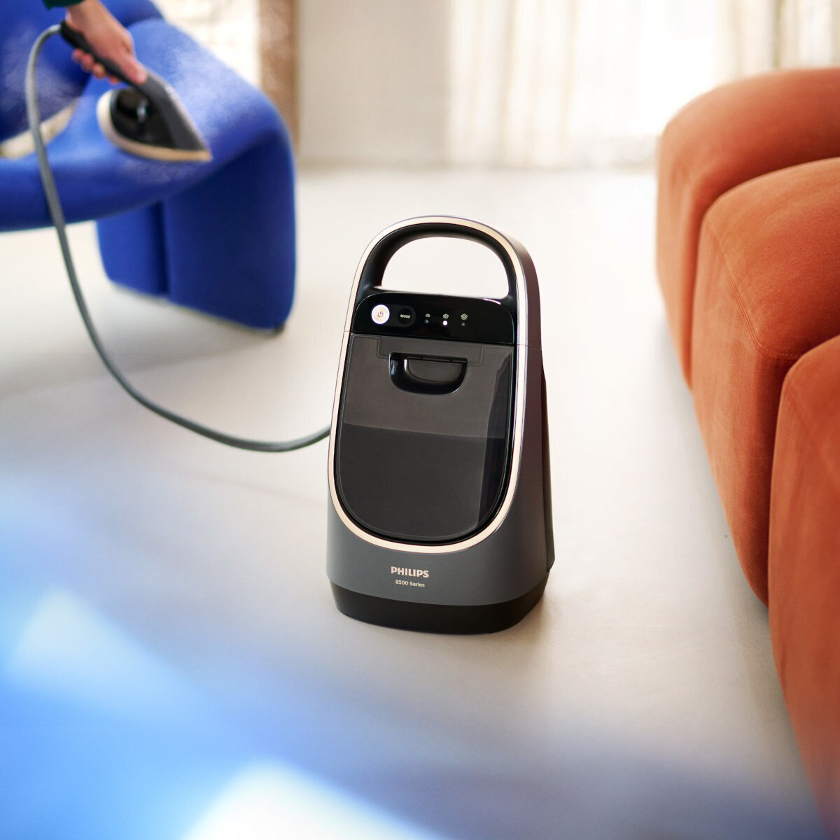 Szara i czarna parownica Philips serii 8500 z uchwytem. Osoba używa jej na niebieskim krześle.