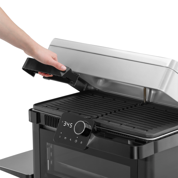 Egy kéz kinyitja a grillt. A grill fekete, digitális időzítővel és rozsdamentes fedéllel.