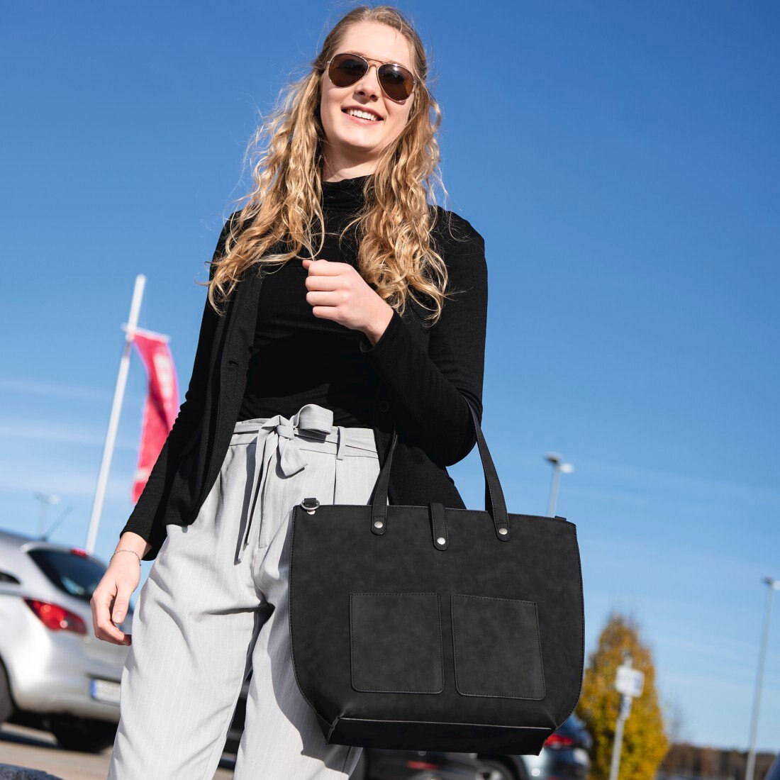 Kobieta w okularach przeciwsłonecznych i czarnej torbie stojąca przed samochodami.