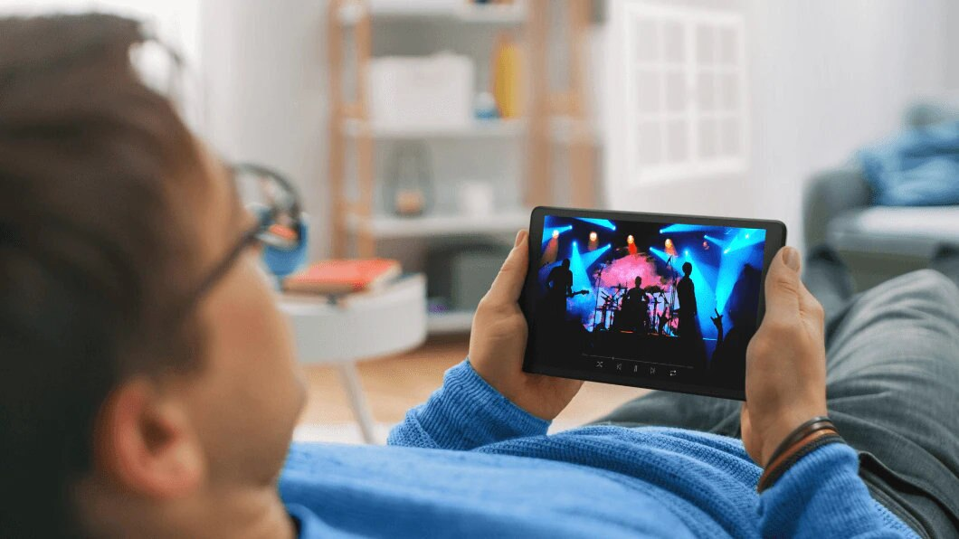 Mężczyzna ogląda wideo z koncertu na tablecie. Siedzi na kanapie.