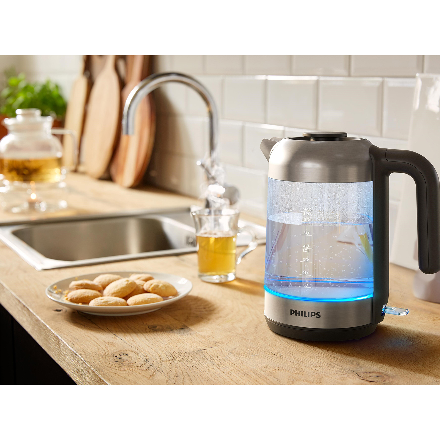 Bollitore elettrico in acciaio inox con luce blu, bicchiere di tè, biscotti e piano di lavoro in legno.