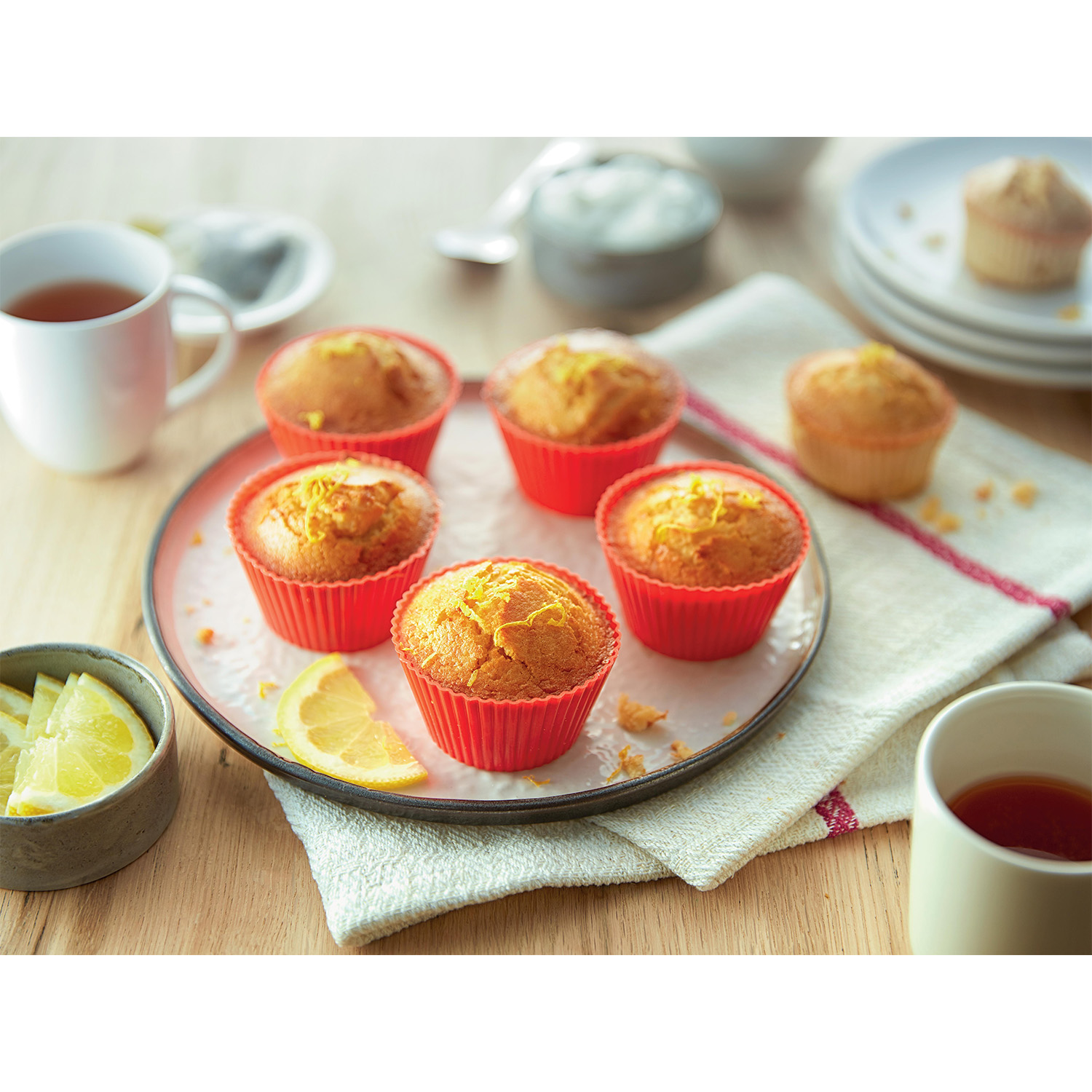 Piatto con muffin in tazze rosse, fette di limone e tazze da tè su un tavolo di legno.