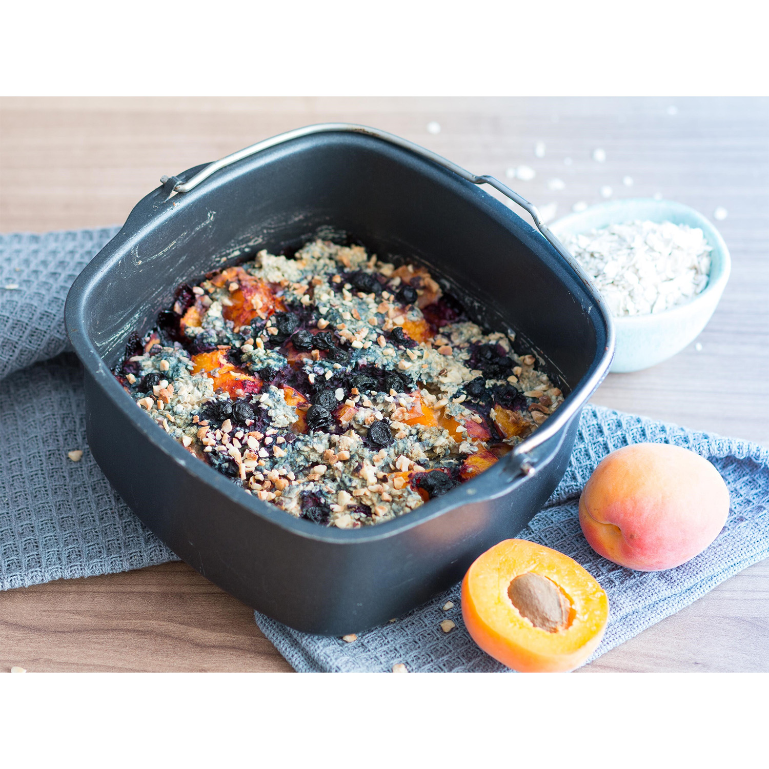 Fiocchi d'avena al forno in padella nera con frutta, ciotola di avena, albicocche su stoffa blu.