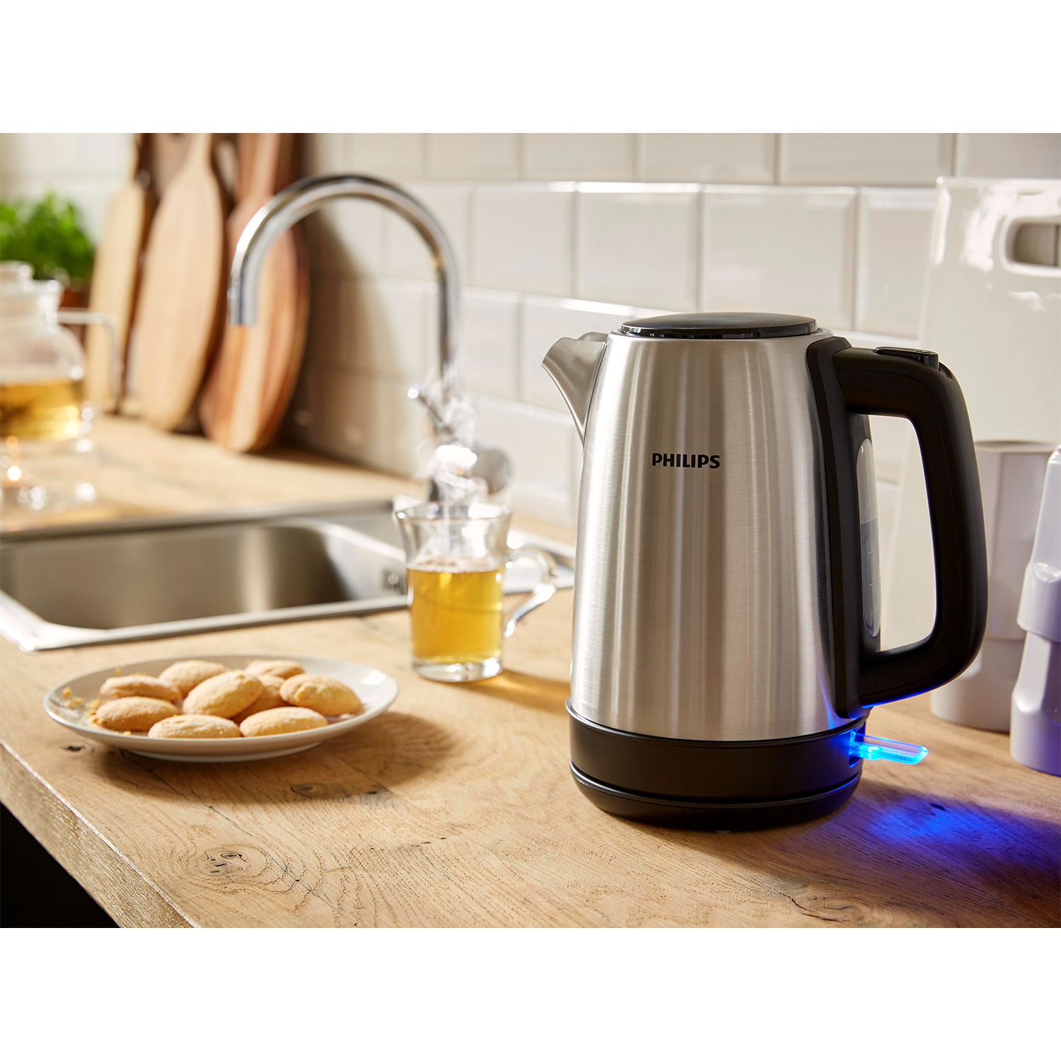 Bollitore elettrico Philips in acciaio inox su un piano di lavoro in legno, acqua versata in un bicchiere. Biscotti sul piatto.