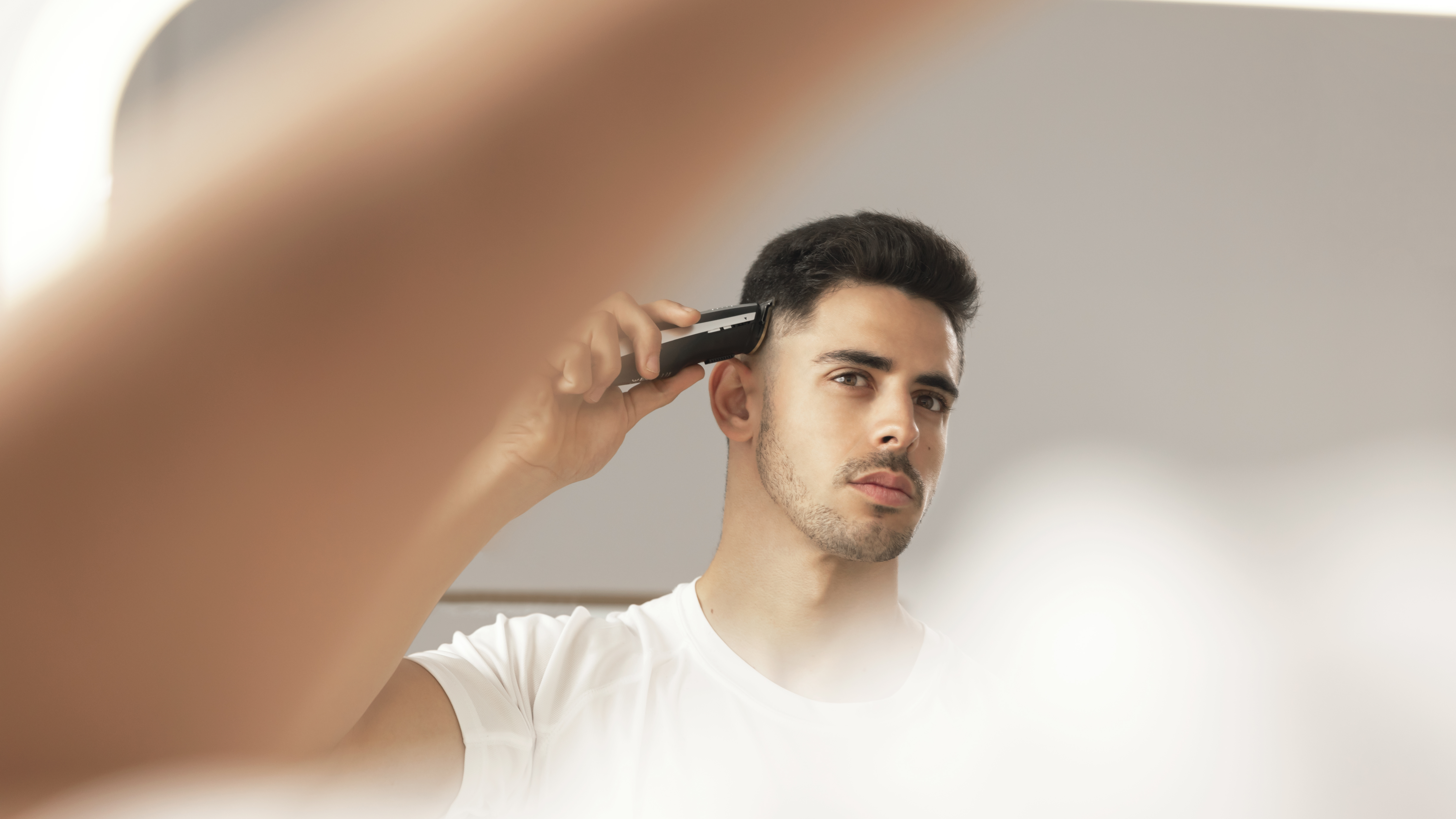 Un hombre en camisa blanca se afeita las patillas con una recortadora eléctrica, mirándose al espejo.