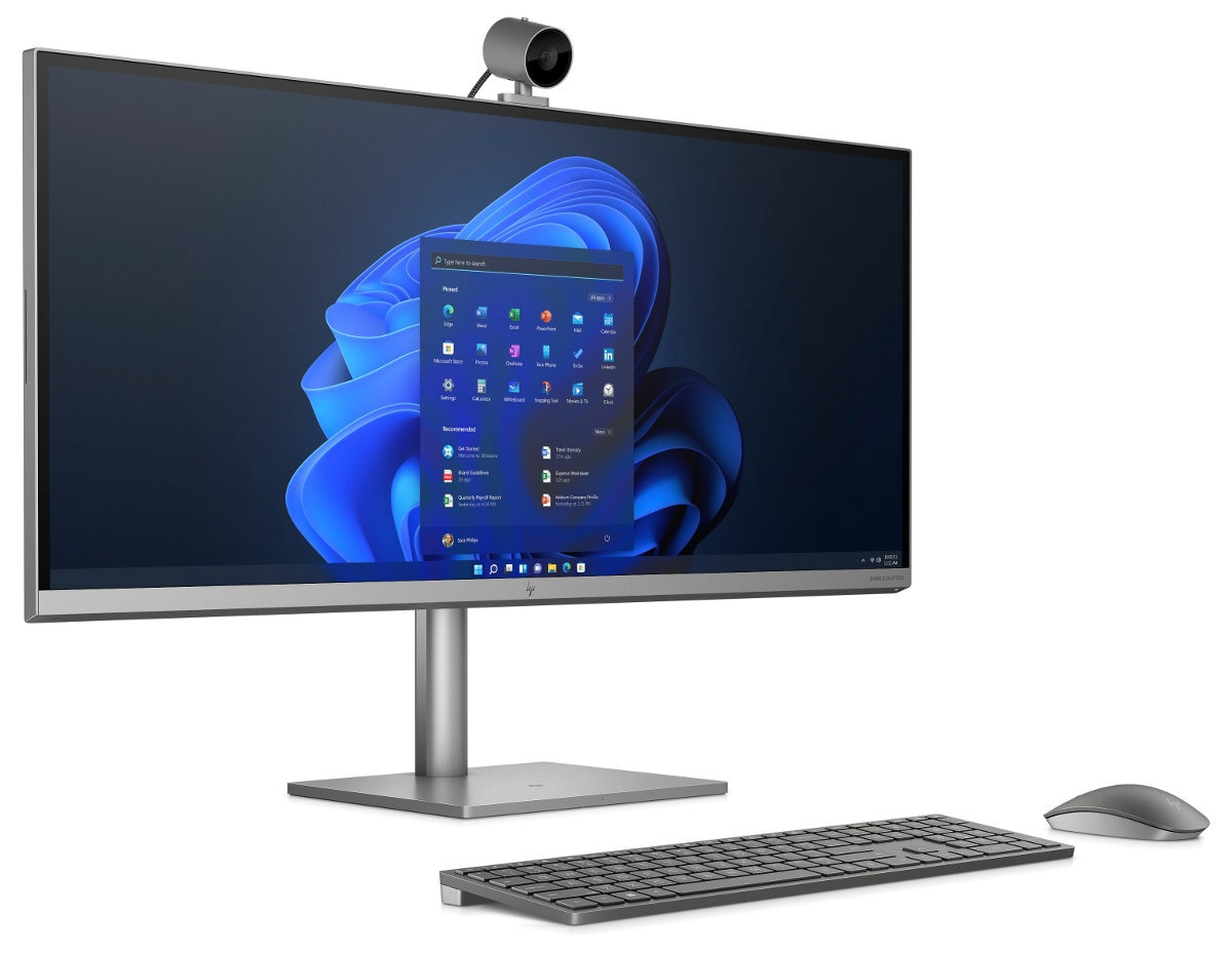 Srebrny monitor z kamerą internetową, klawiaturą i myszą na białej powierzchni. Ekran Windows 11.