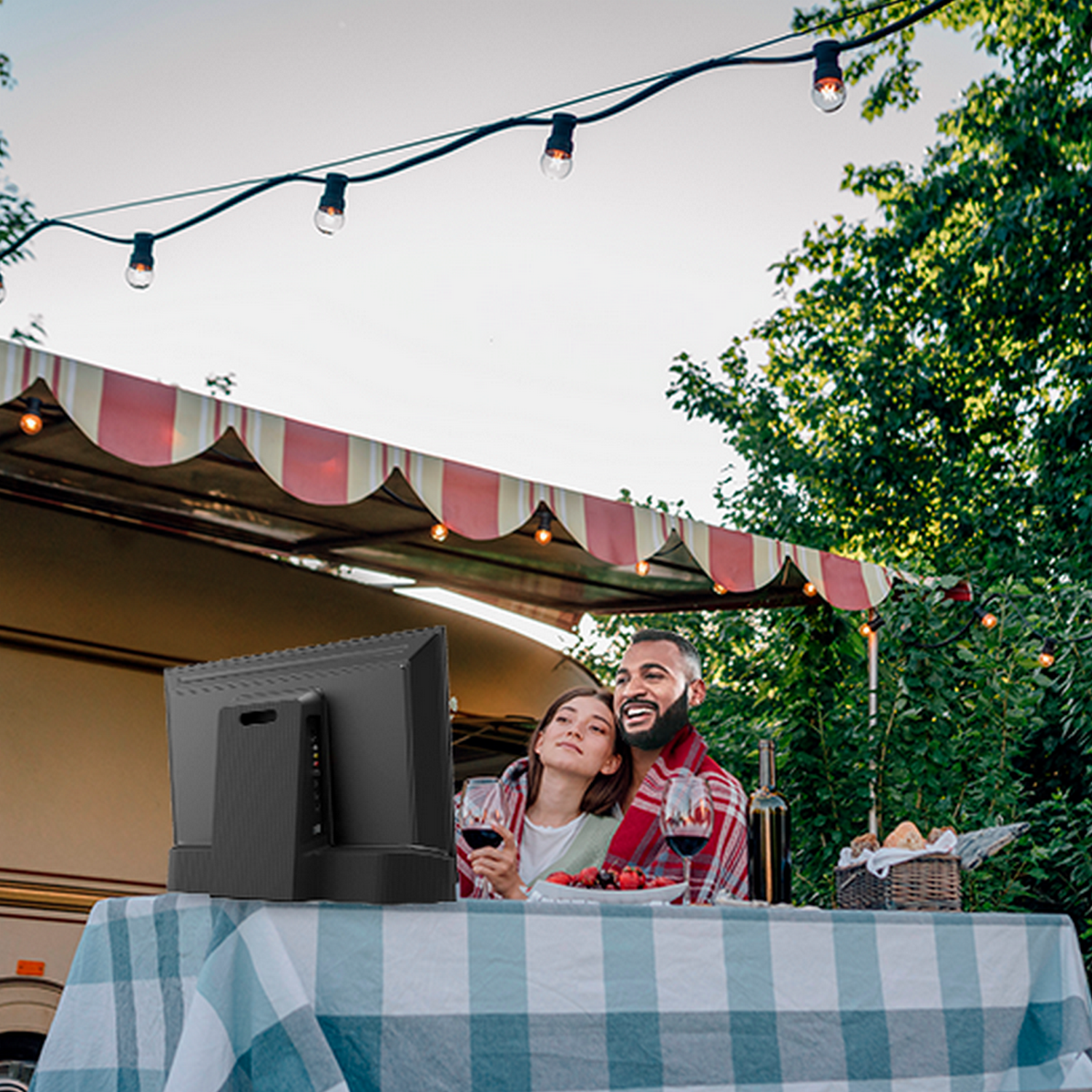 Koppel geniet van wijn aan een tafel buiten een camper. Lichten erboven, bomen op de achtergrond.