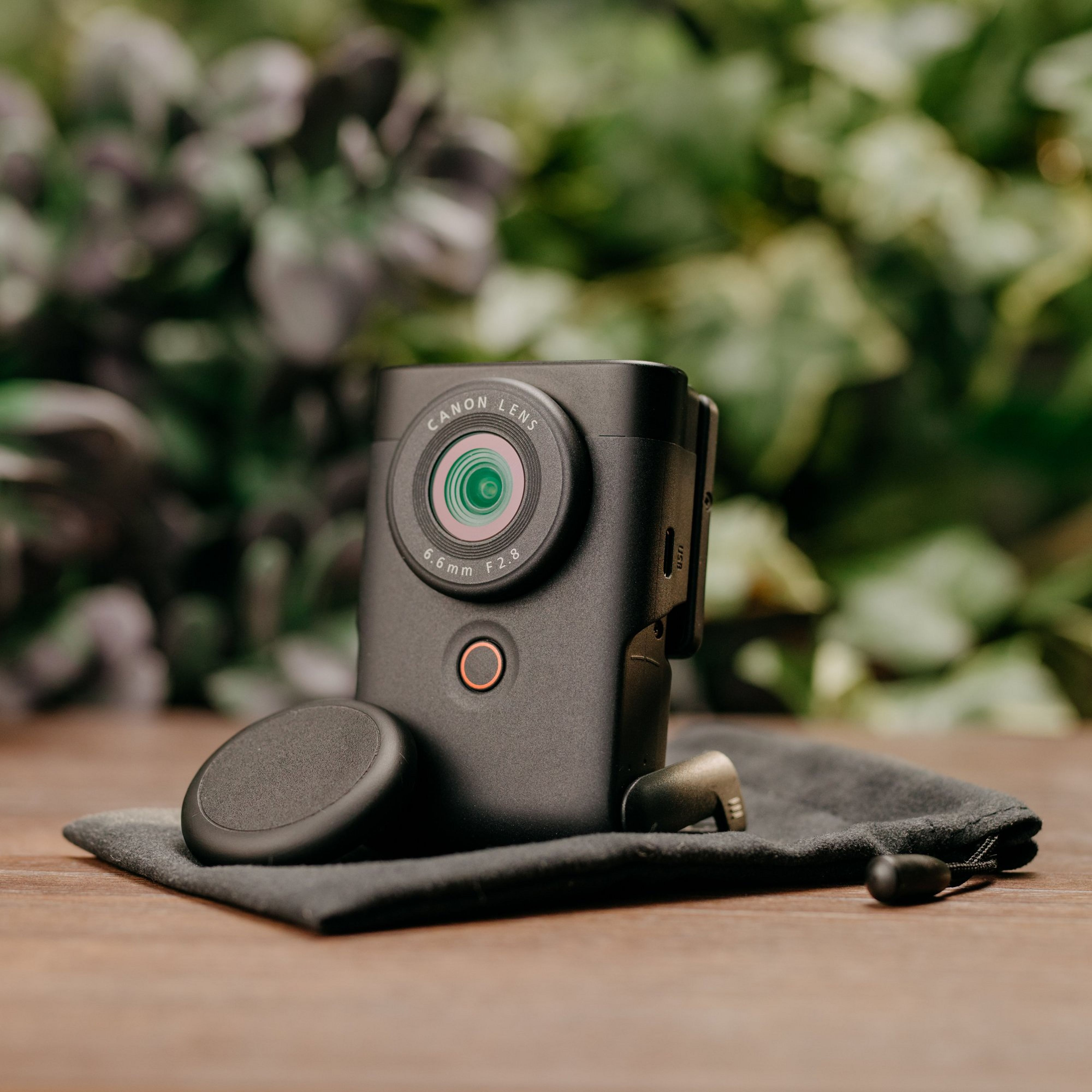 Un appareil photo Canon noir avec un objectif, un bouchon magnétique et une pochette. Feuillage vert en arrière-plan.
