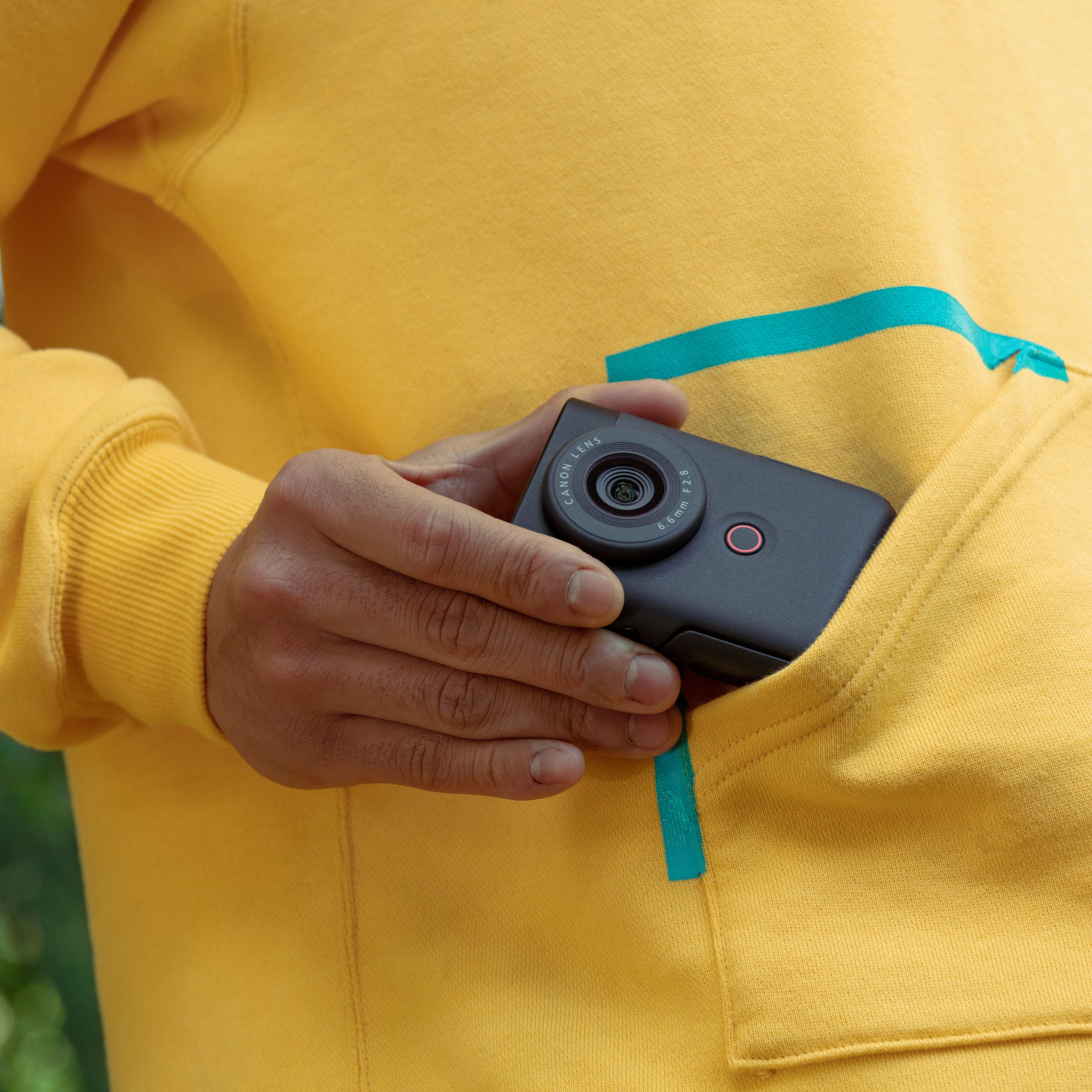 Une personne tient un appareil photo Canon noir dans la poche d'un sweat à capuche jaune. La poche a une bordure turquoise.
