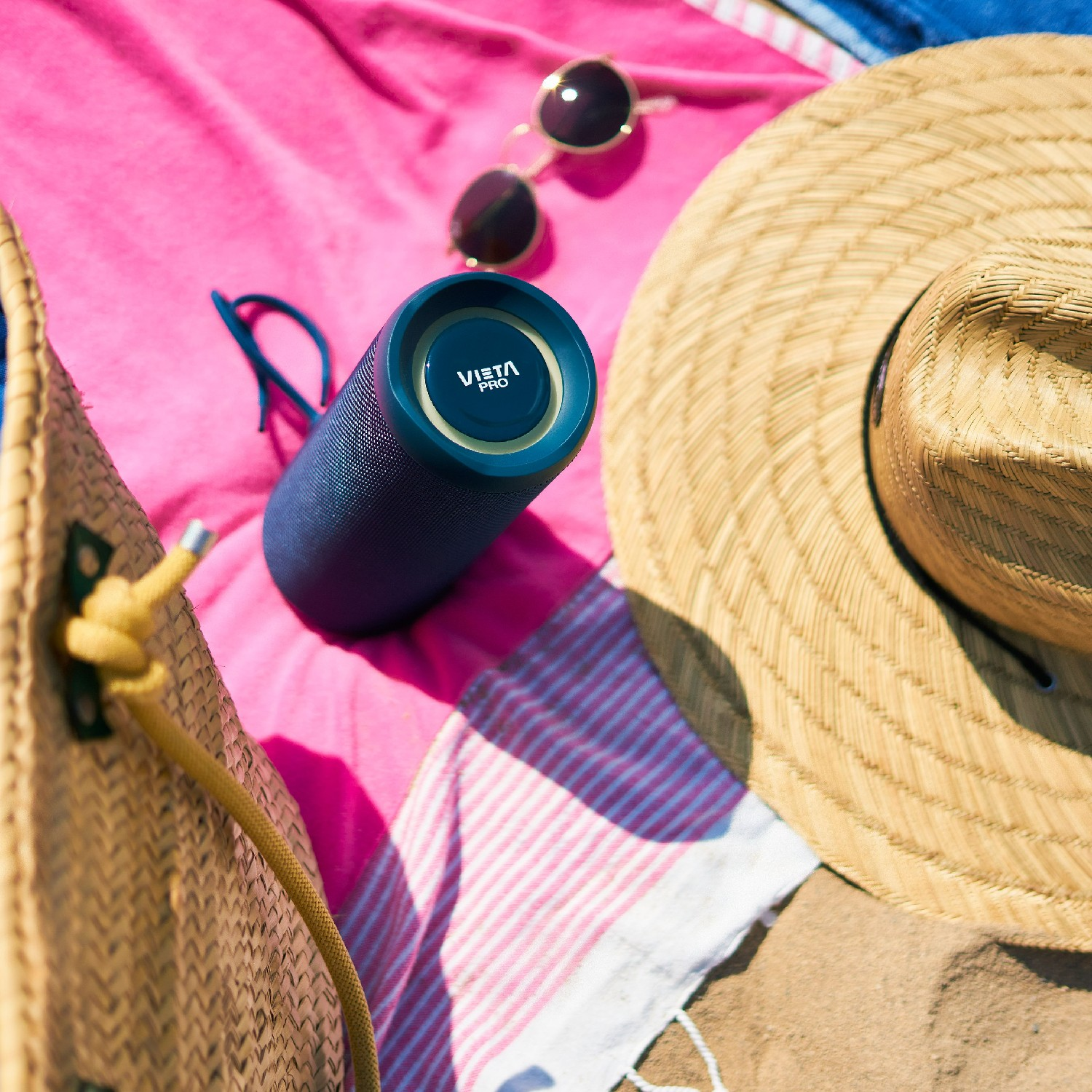 Altavoz azul en una toalla rosa con sombrero de paja, gafas de sol y bolso de paja en la playa.