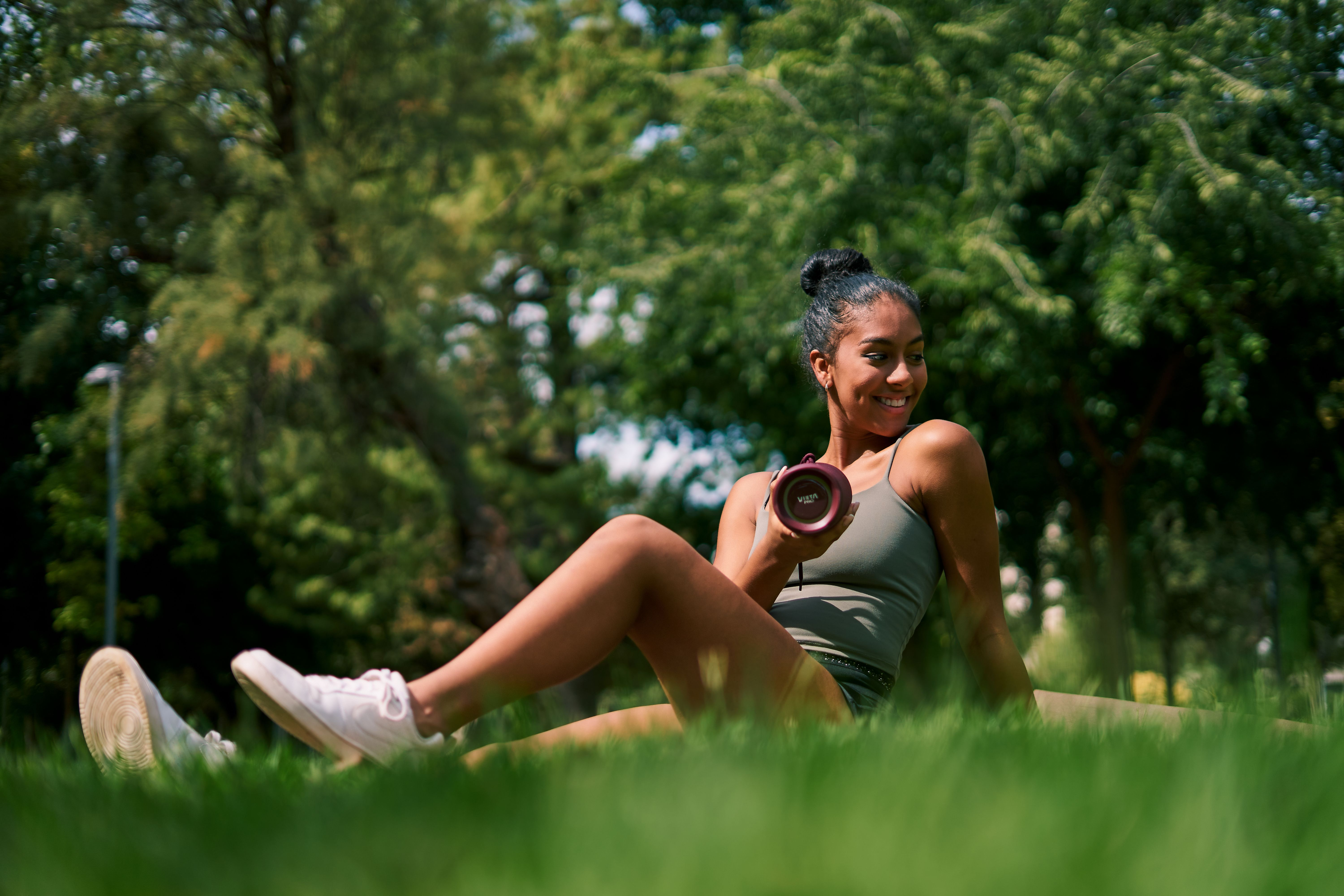 Mujer en el parque, sosteniendo un altavoz, sonriendo. Hierba verde y árboles al fondo.