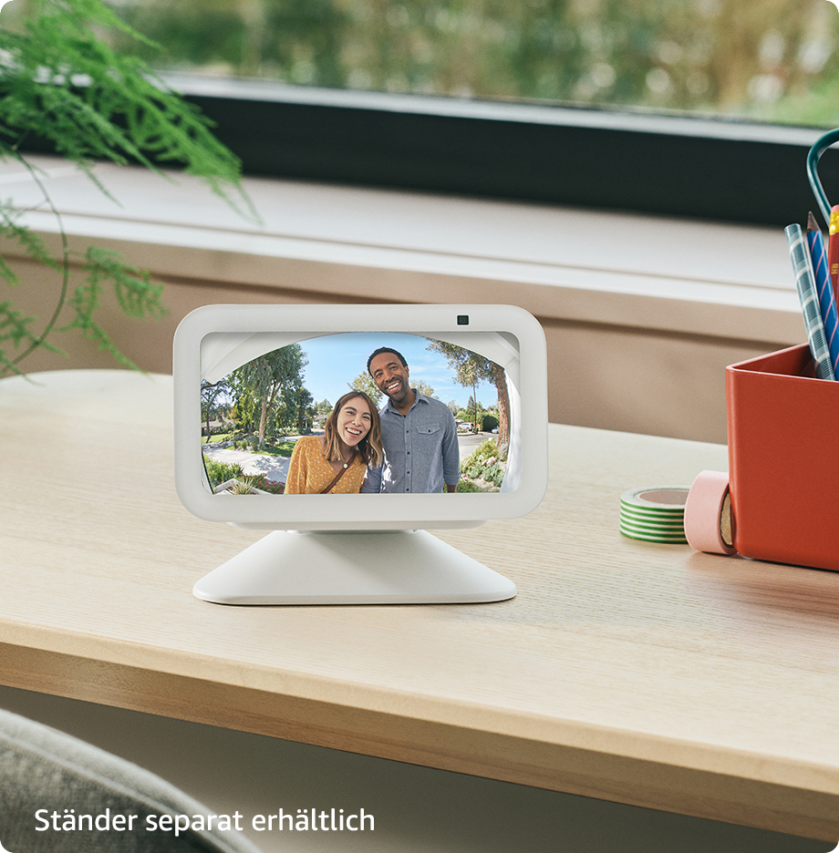 Weißes Smart Display zeigt lächelndes Paar. Das Display steht auf hellem Holz nahe einem Fenster.