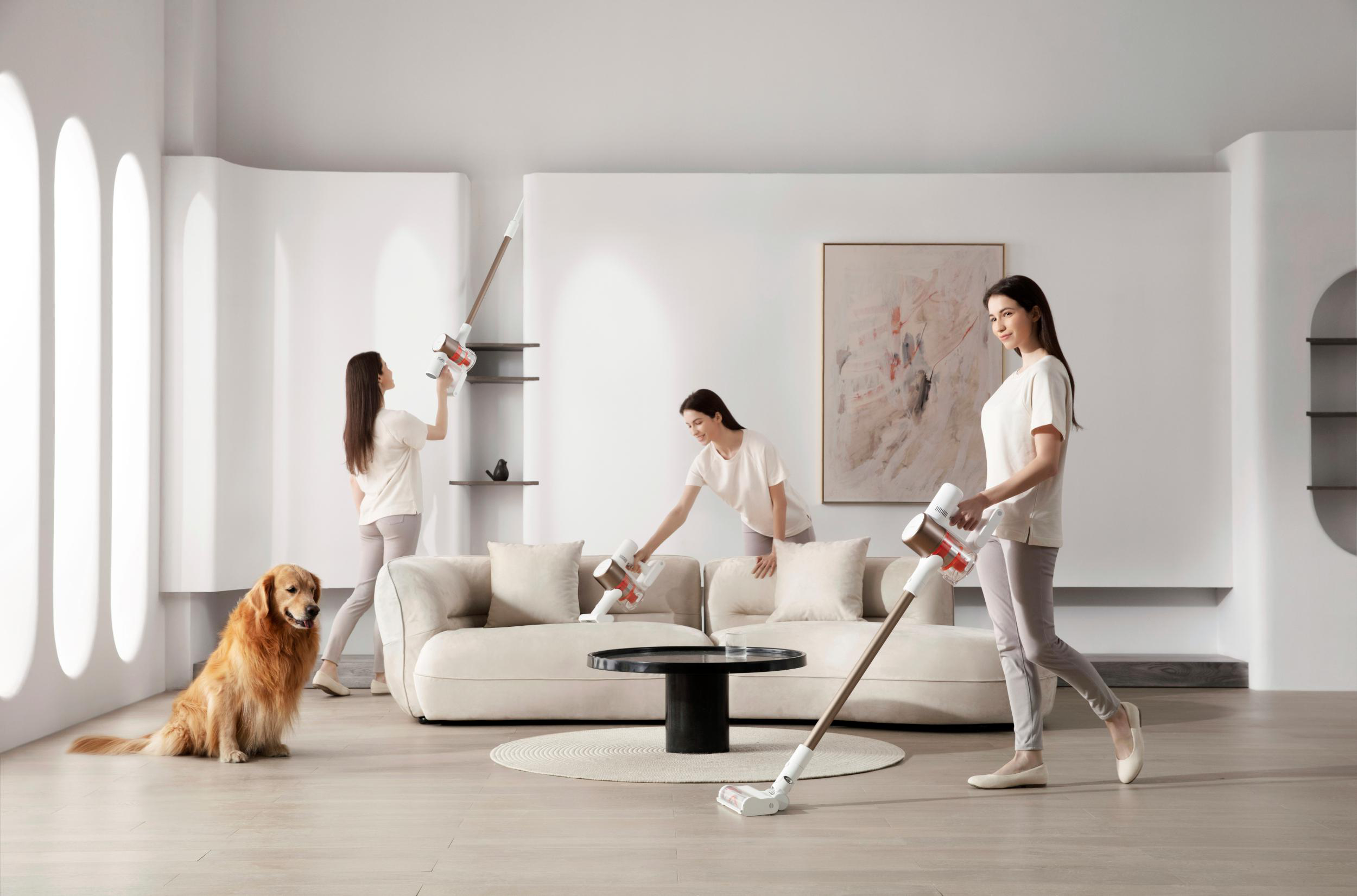 Drei Frauen benutzen einen kabellosen Staubsauger in einem Wohnzimmer mit einem Hund. Der Raum ist hell und modern.