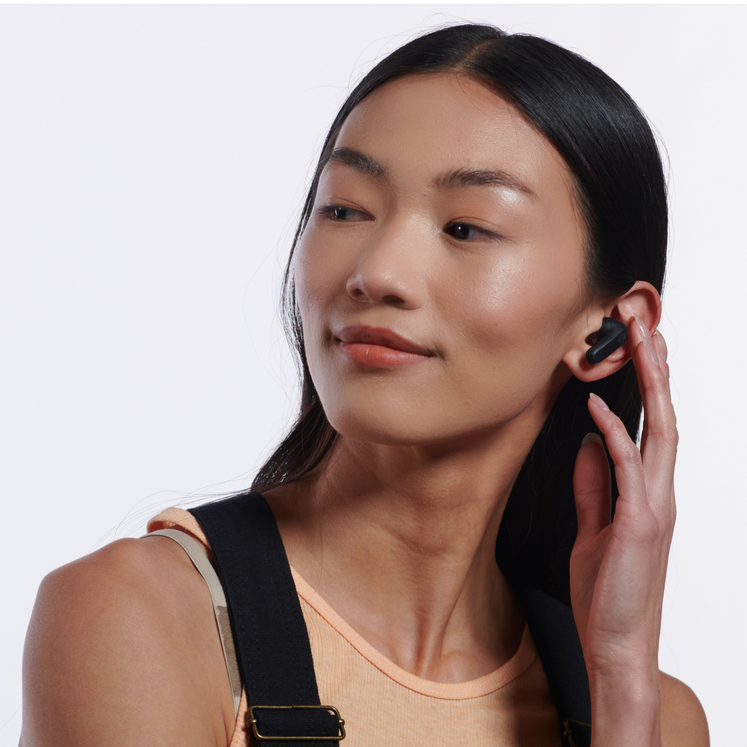 Woman wearing black wireless earbuds in her left ear, touching her ear, white background.