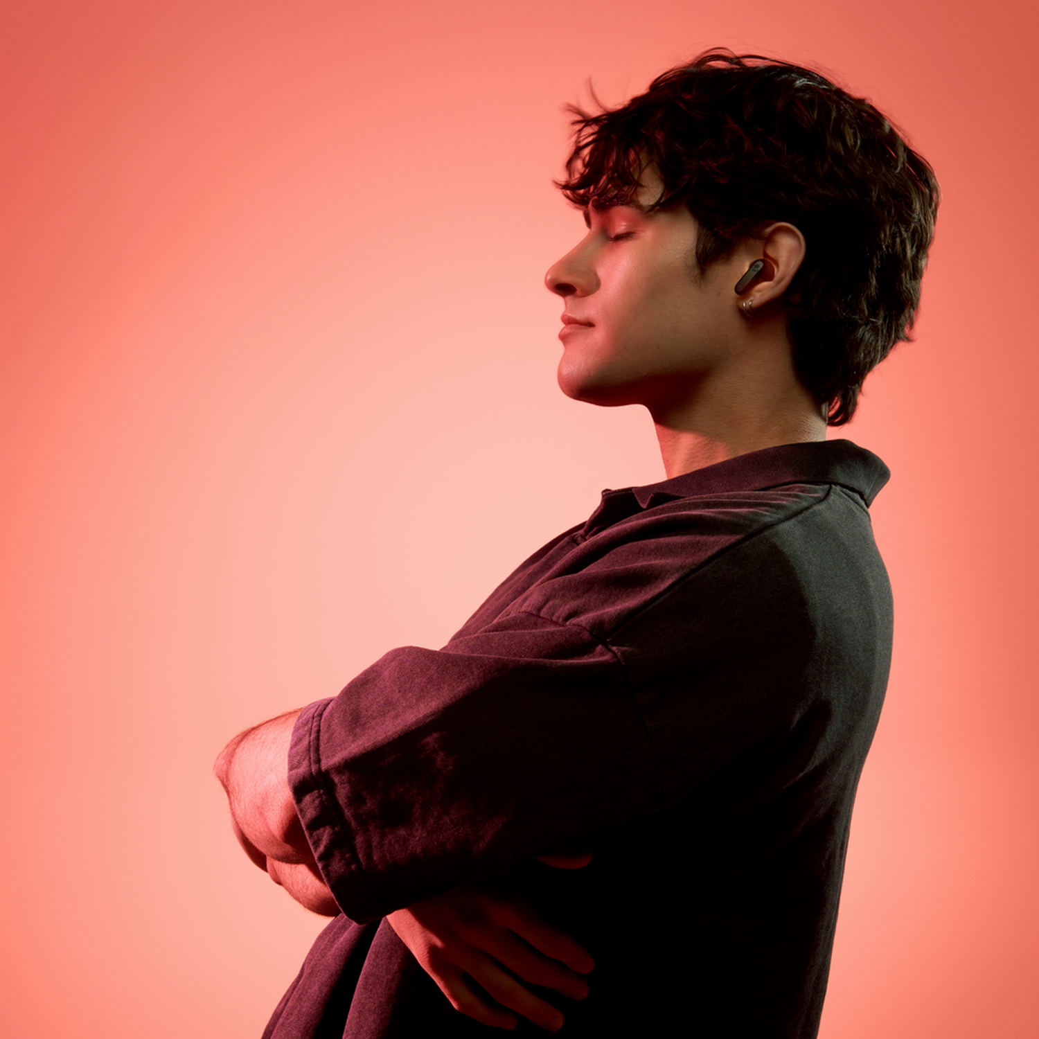 A man with closed eyes, arms crossed. He wears a dark shirt against a red background.
