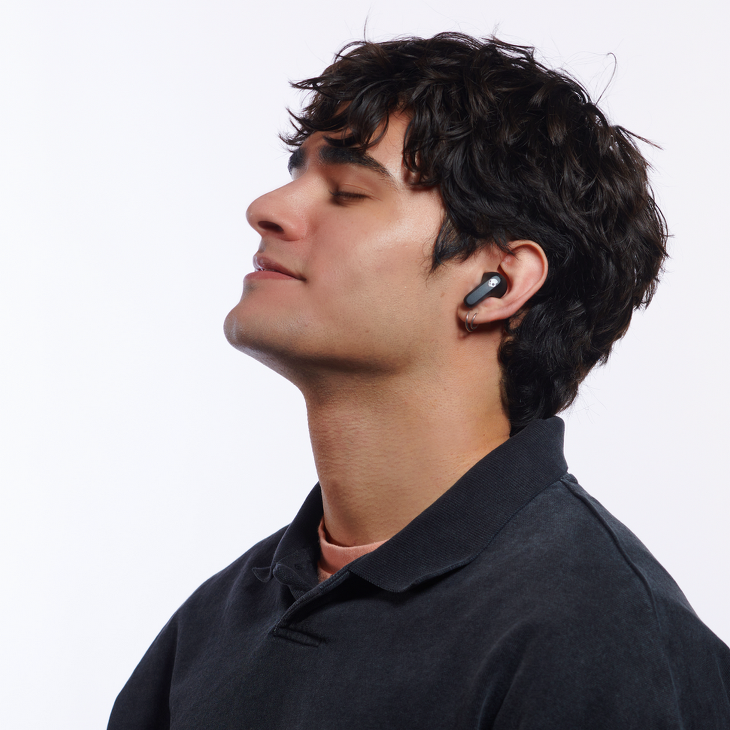 Man with closed eyes wearing black earbuds. Light skin, dark hair, dark shirt. White background.