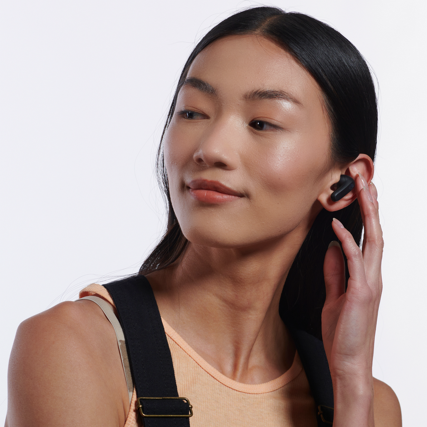 Woman with black earbud in ear, wearing orange top and black overalls. Smiling, looking away.