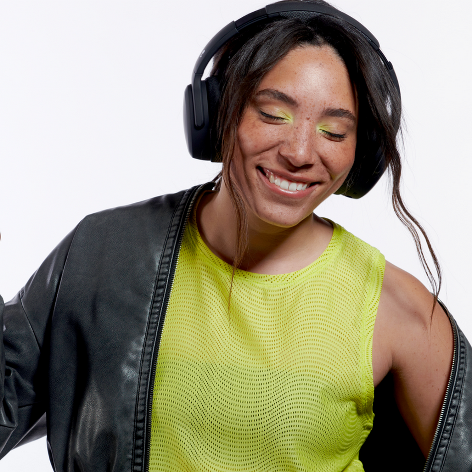 Woman smiling with black headphones and a neon green shirt and black jacket. Yellow eye makeup.