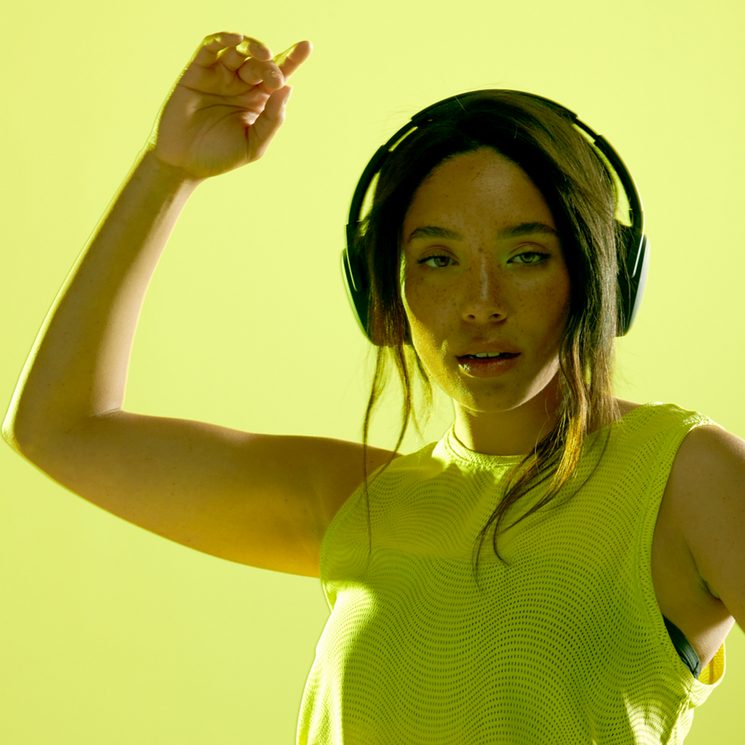 Woman wearing headphones, raising arm, and looking at the camera with a green background.