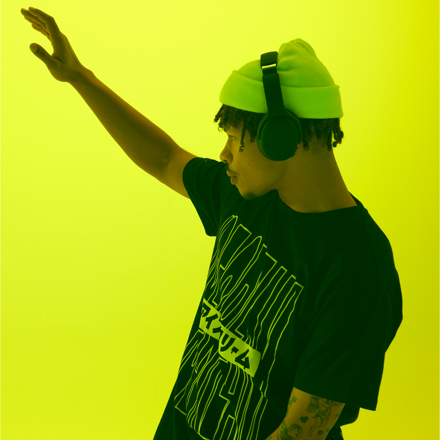 Young man with green beanie and headphones. He is raising one arm up. Yellow background.