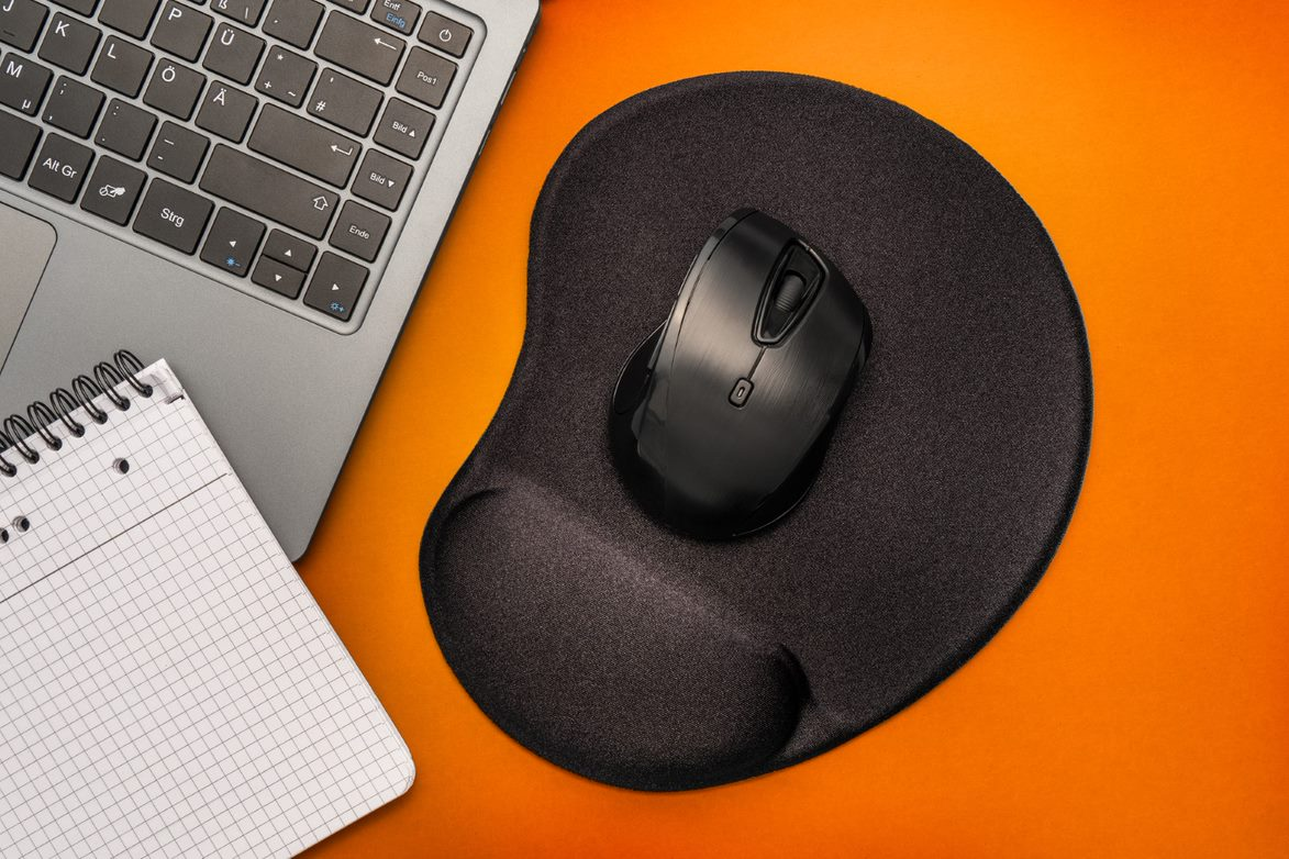 Ein Laptop, eine Maus und ein Notizblock auf orangefarbenem Hintergrund. Schwarzes Mauspad.