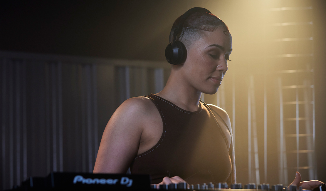 DJ femme à une table de mixage, portant un casque. Cadre sombre avec lumières.
