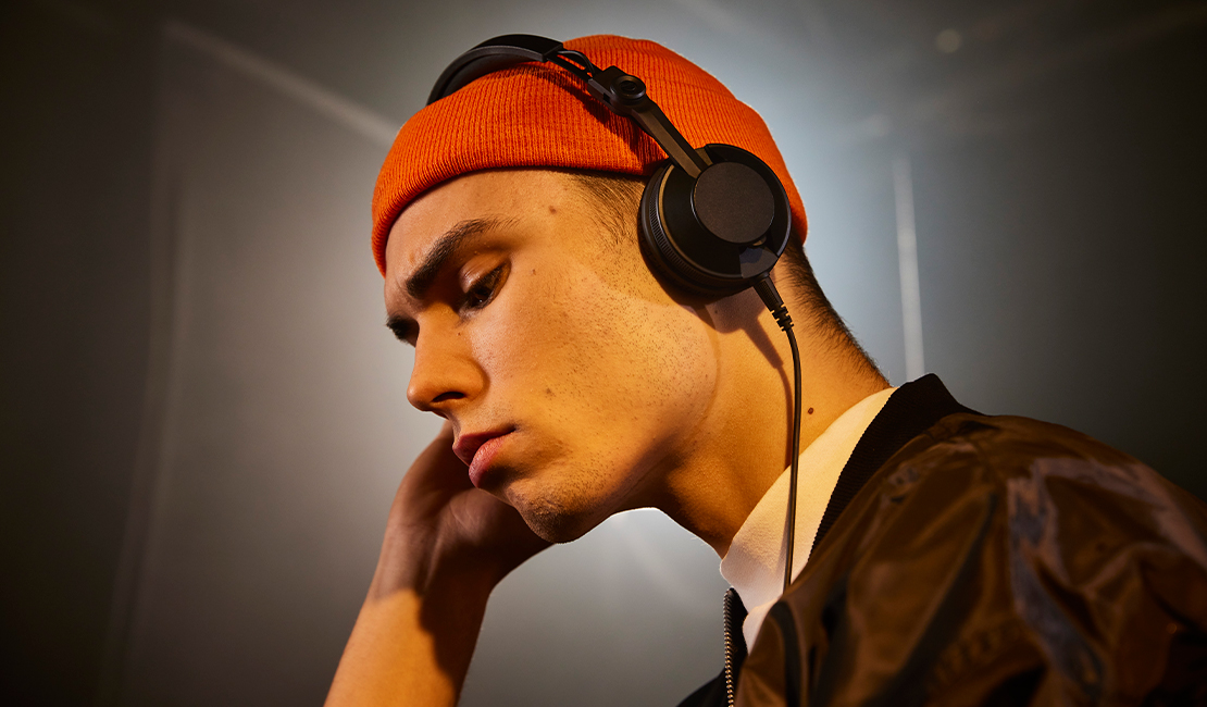 Un jeune homme portant un bonnet orange et un casque noir. Il regarde vers le bas. Il porte une veste noire sur une chemise blanche.