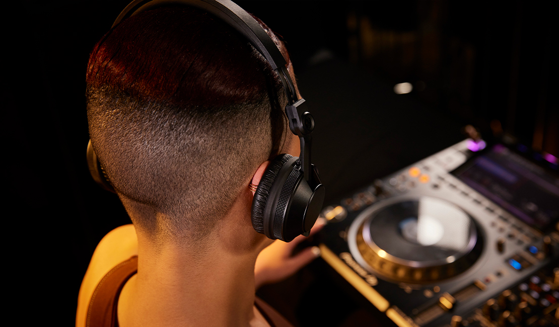 Une personne portant un casque regarde une table de mixage DJ. La personne a les cheveux courts avec des mèches rouges.